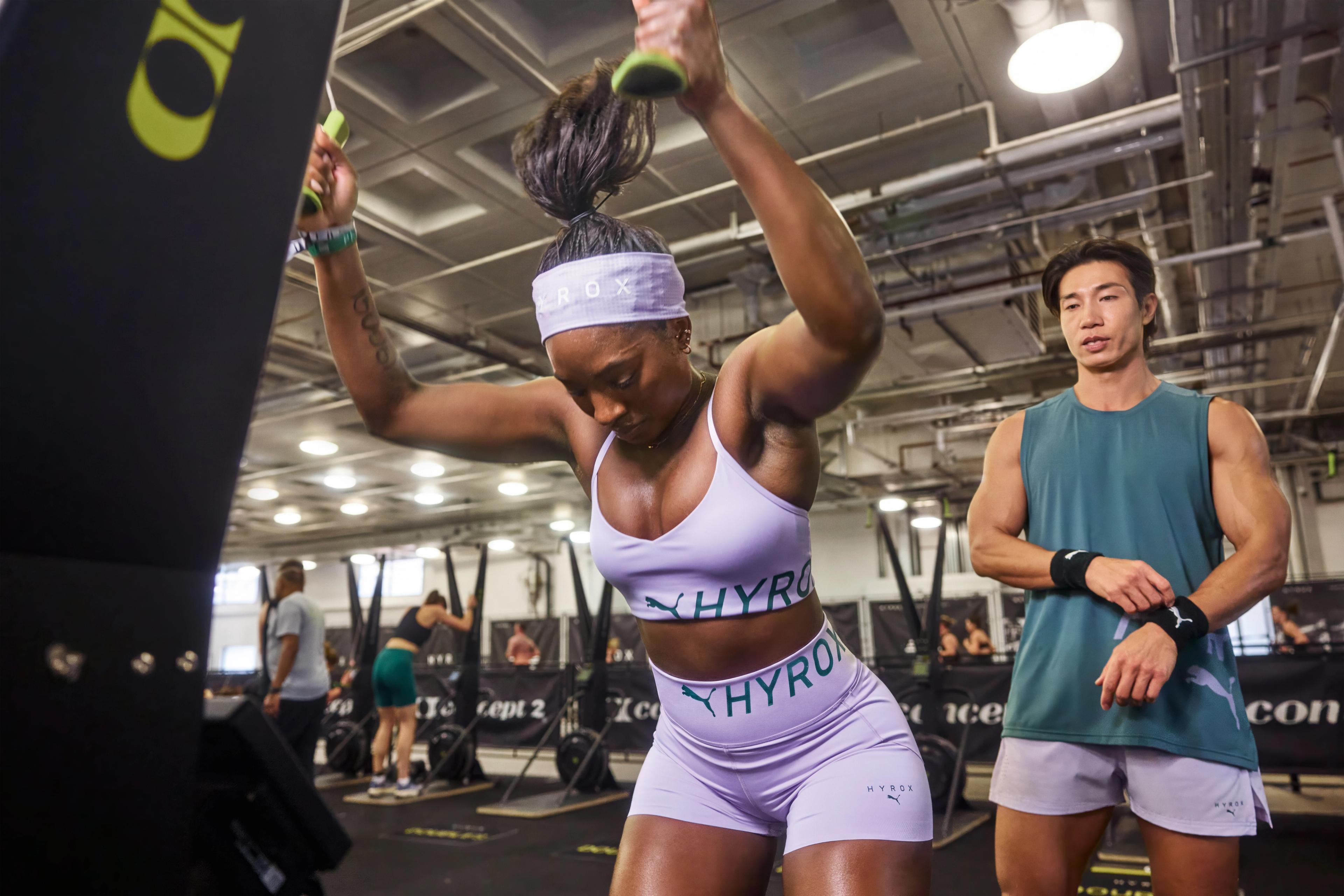 A woman in a purple HYROX athletic outfit pulls down handles on a fitness machine while a man in a green tank top watches.