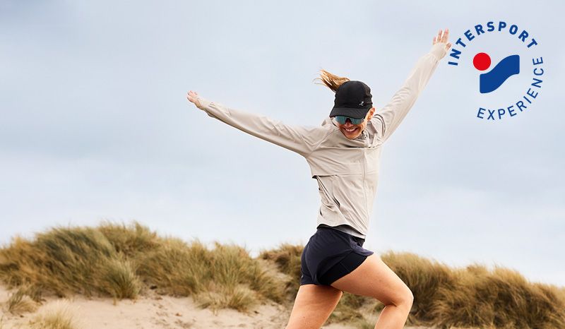 A smiling woman in athletic wear jumps on sandy dunes with the Intersport Experience logo.