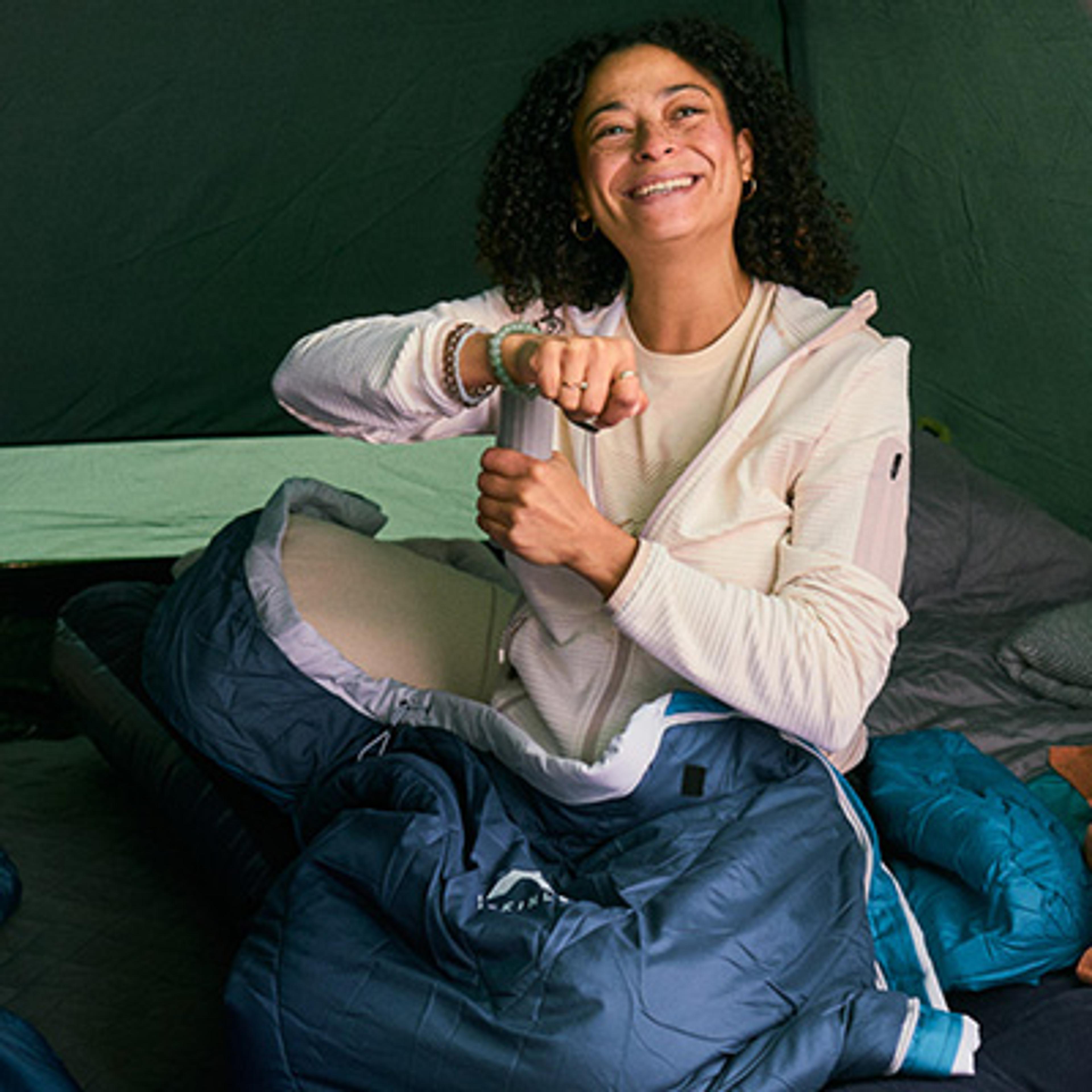 A smiling woman with curly hair sits inside a tent, partially in a blue sleeping bag.