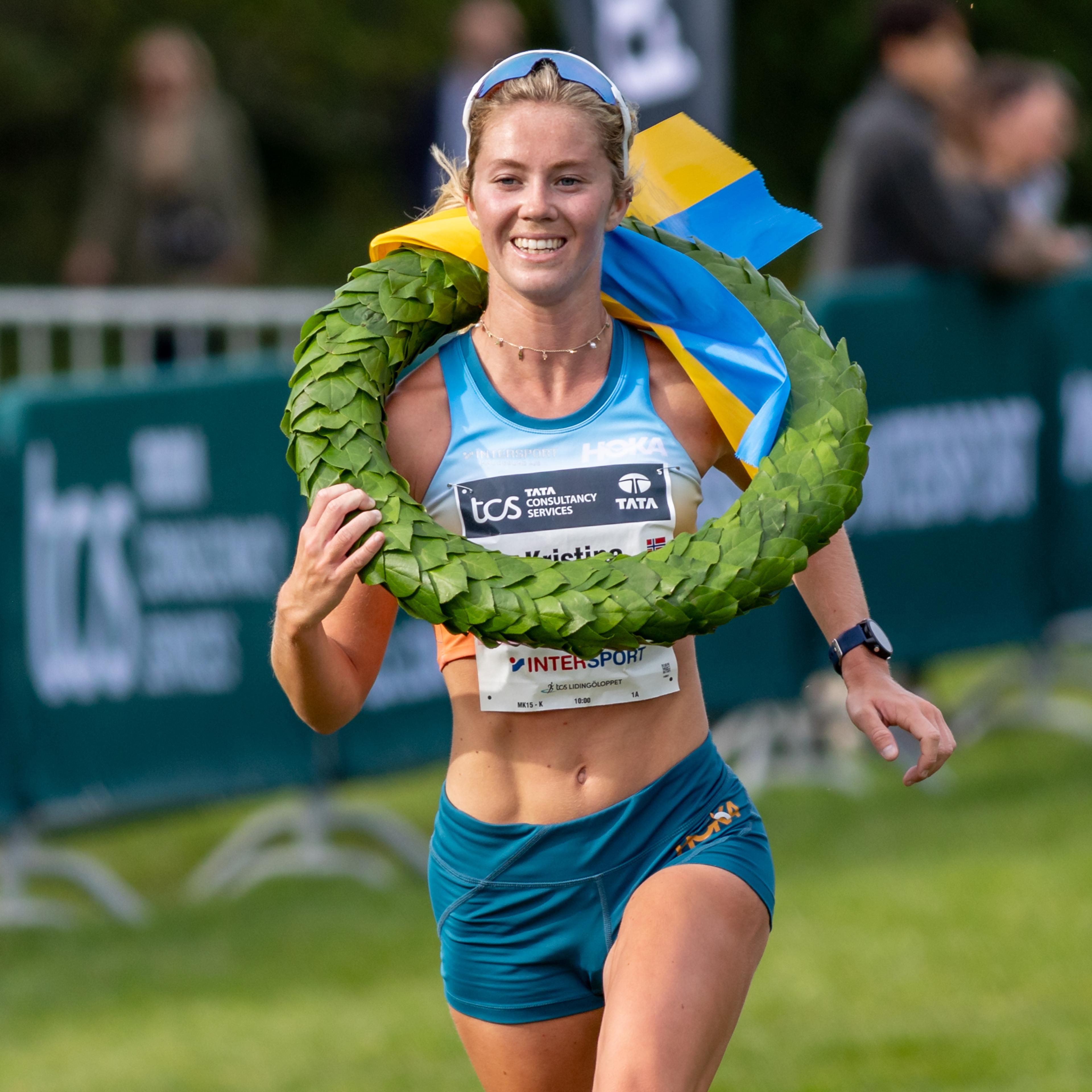 A smiling female athlete with a green wreath and a Ukrainian flag draped over her shoulder at a race.