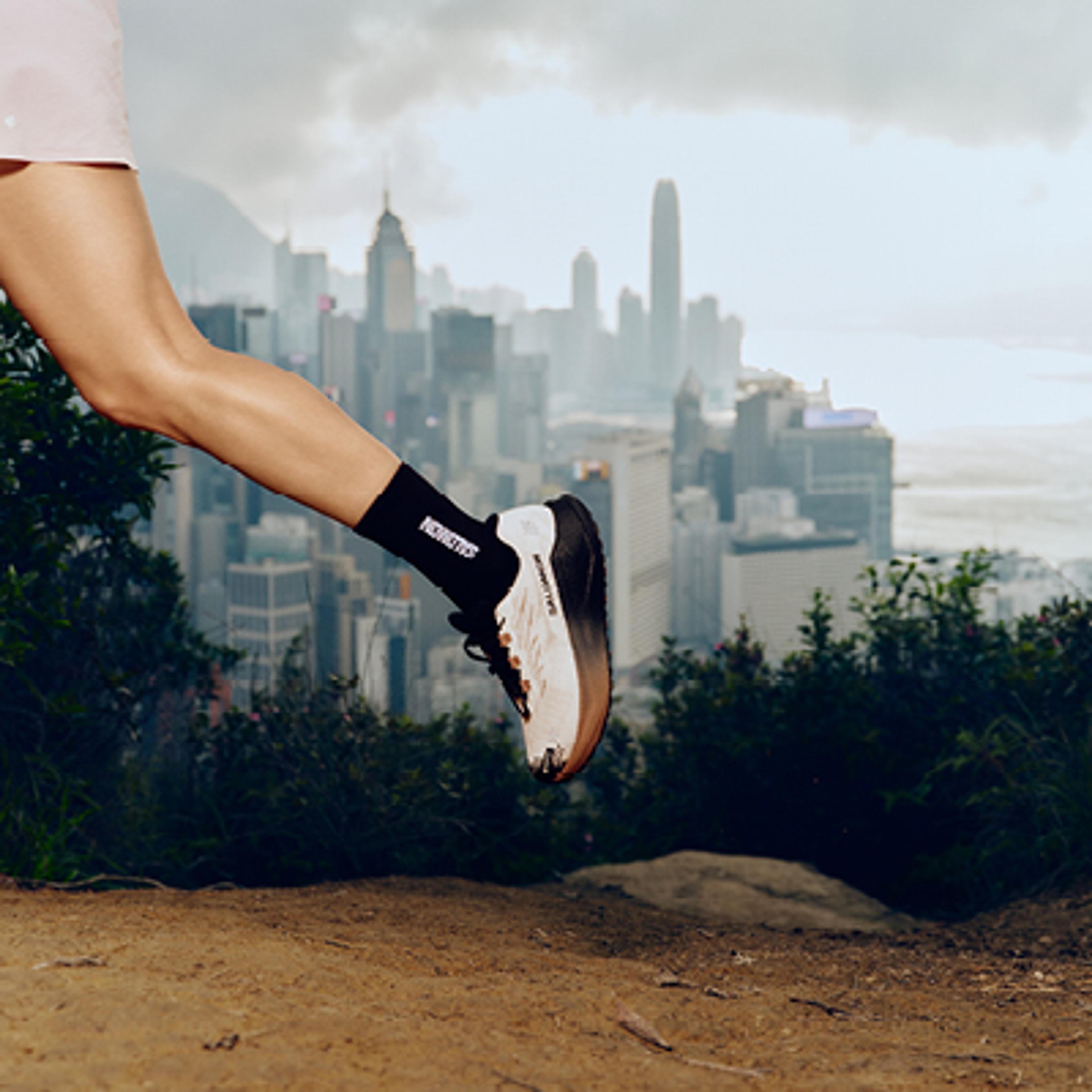 A trail runner's leg and Salomon shoe in mid-stride on a dirt path overlooking a city skyline.