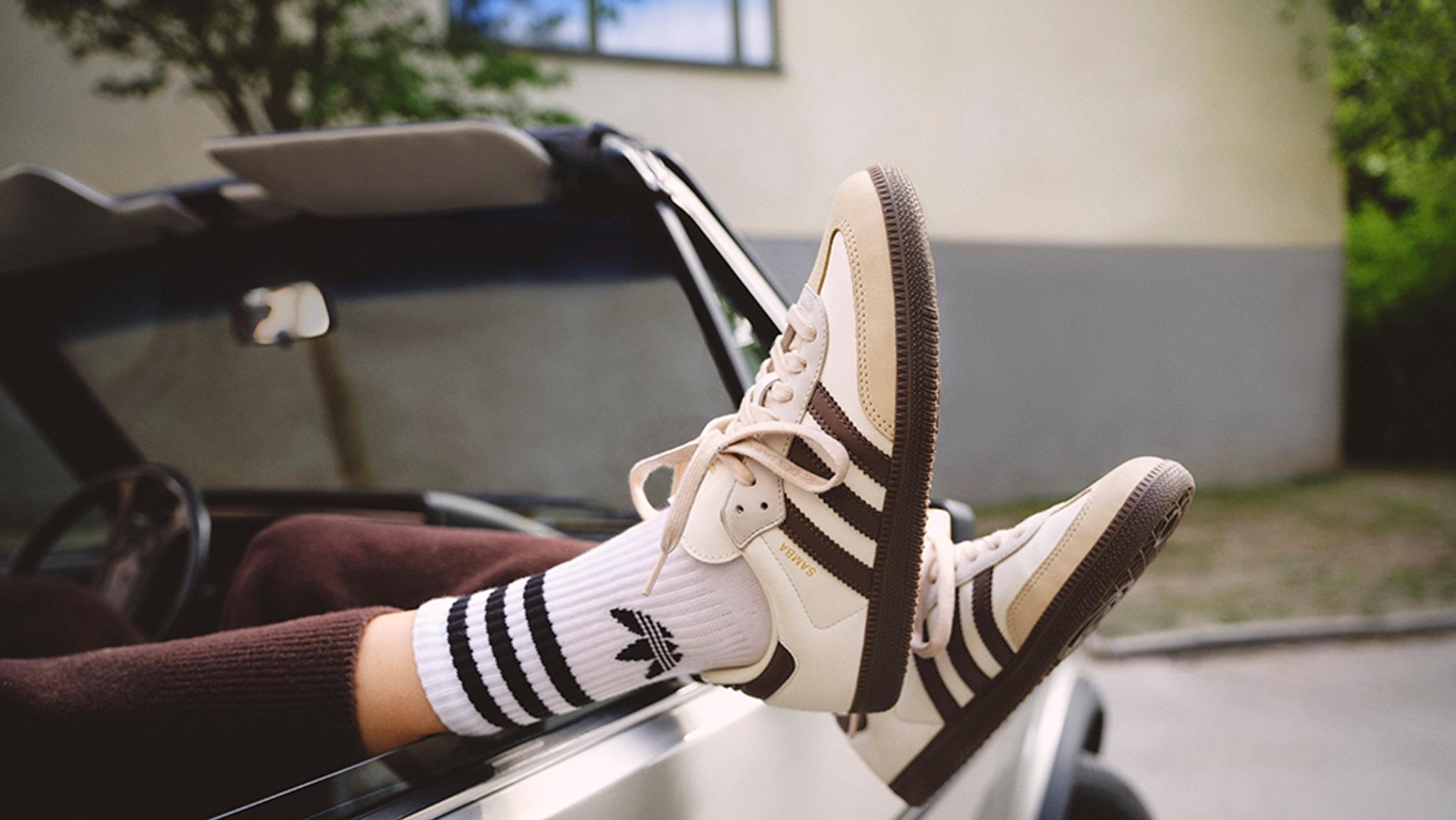 Legs wearing white Adidas socks and beige-brown Samba sneakers propped out of a convertible car window.