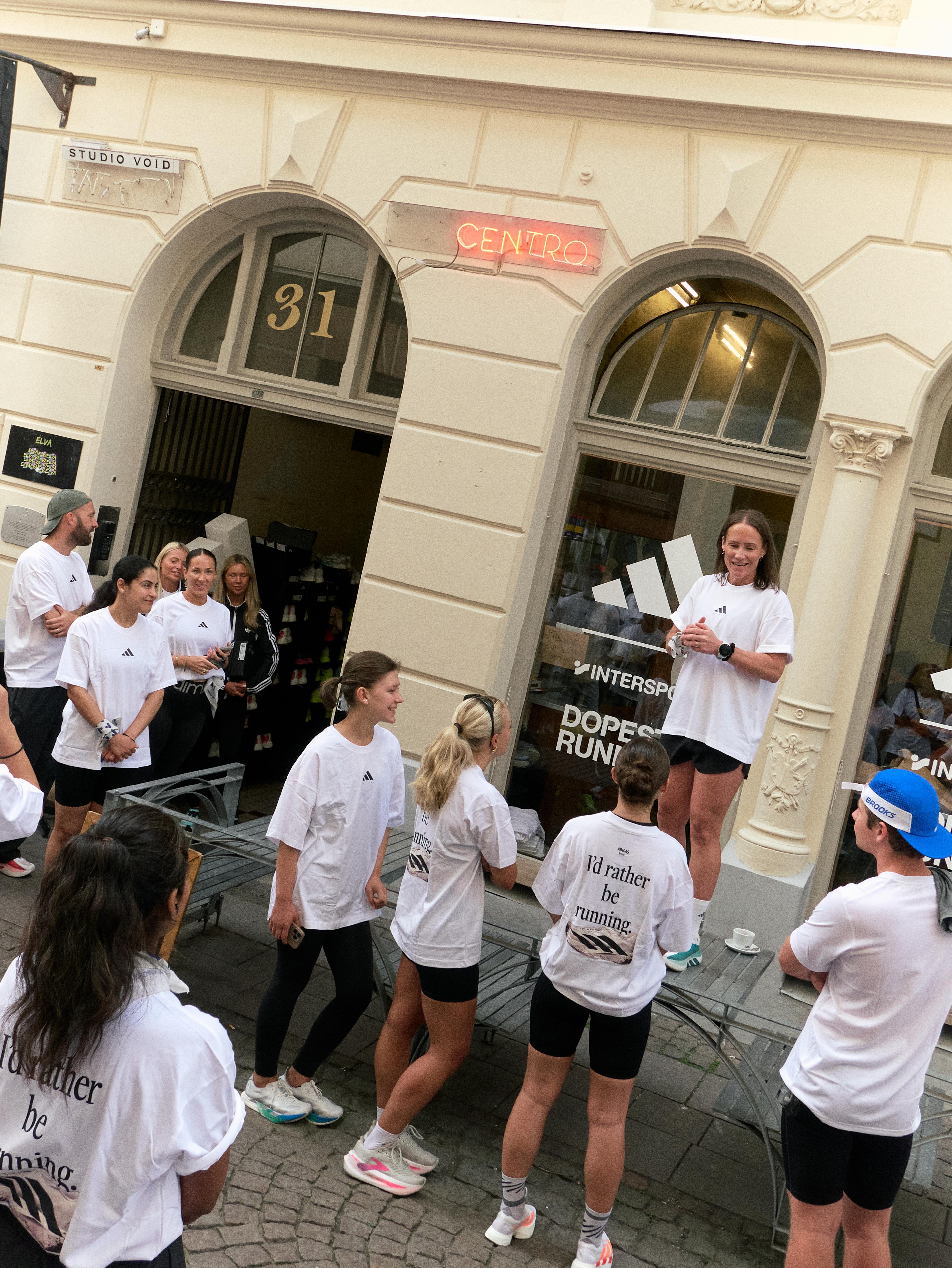 A group of people in white Adidas t-shirts, some reading "I'd rather be running," listen to a woman speaking from a table outside storefronts.