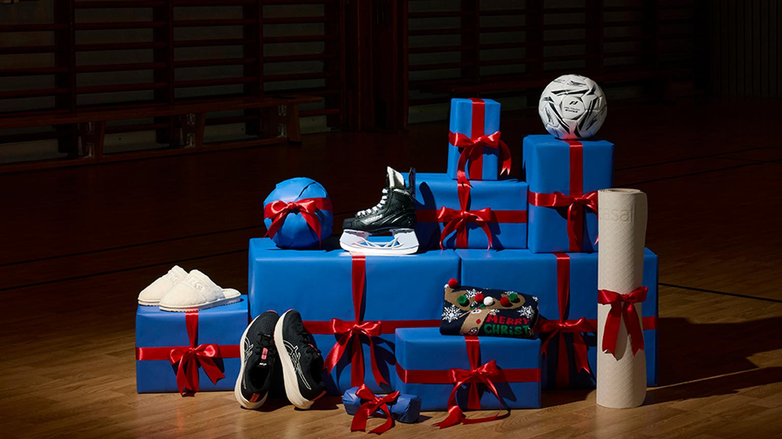 Blue wrapped Christmas gifts with red ribbons, featuring sports equipment like a soccer ball, ice skate, and sneakers, on a gym floor.