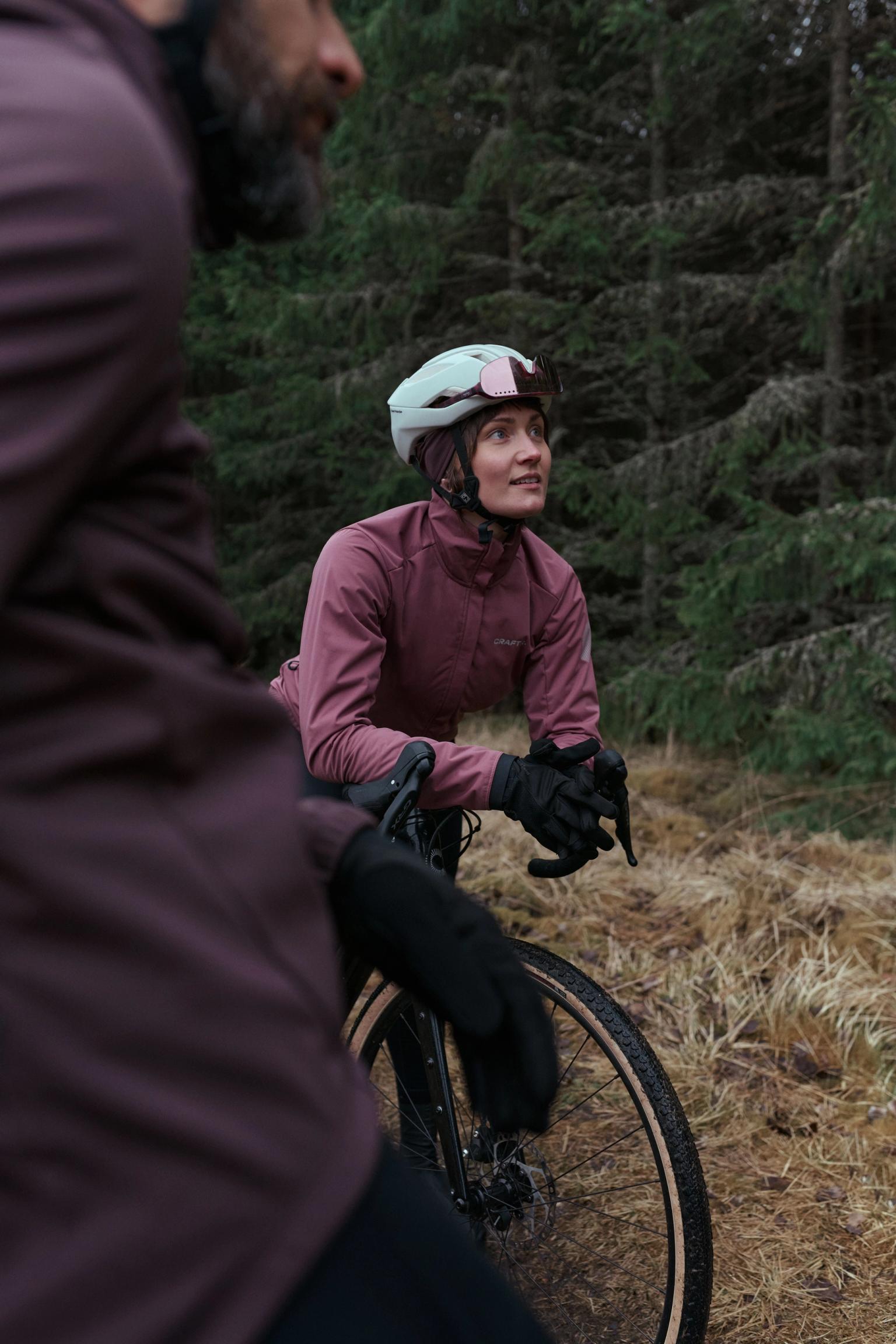 A female cyclist in a pink jacket and helmet looks up from her bike in a forest, with another person blurred in the foreground.