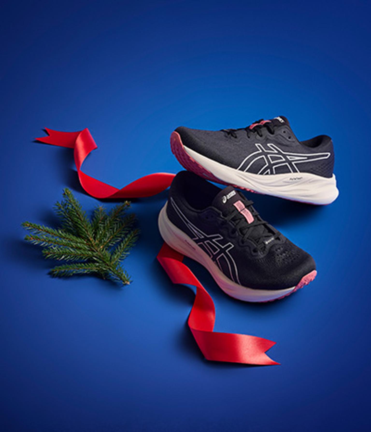 Two black and pink running shoes with a red ribbon and pine sprig on a blue background.