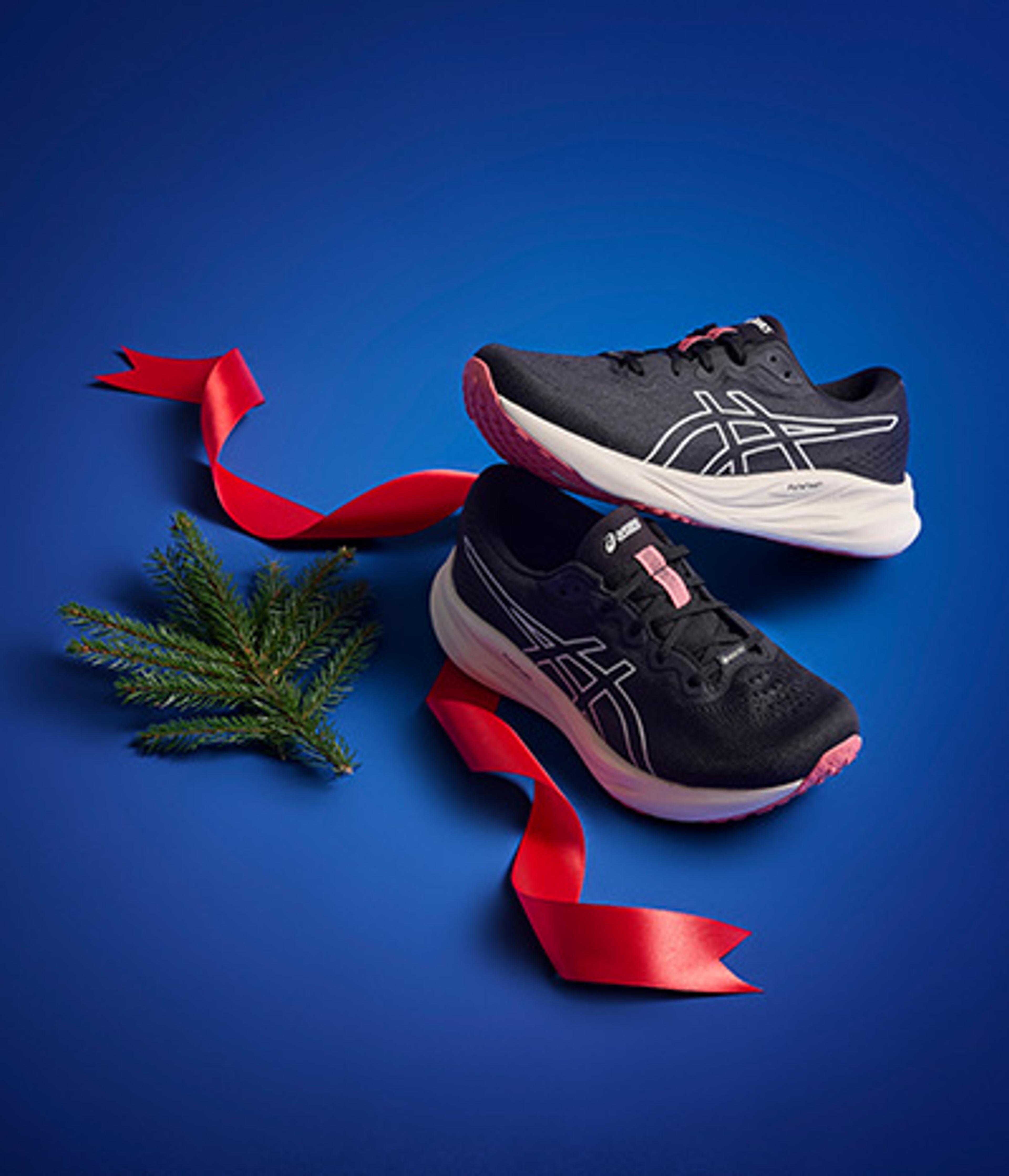 Two black and pink running shoes with a red ribbon and pine sprig on a blue background.