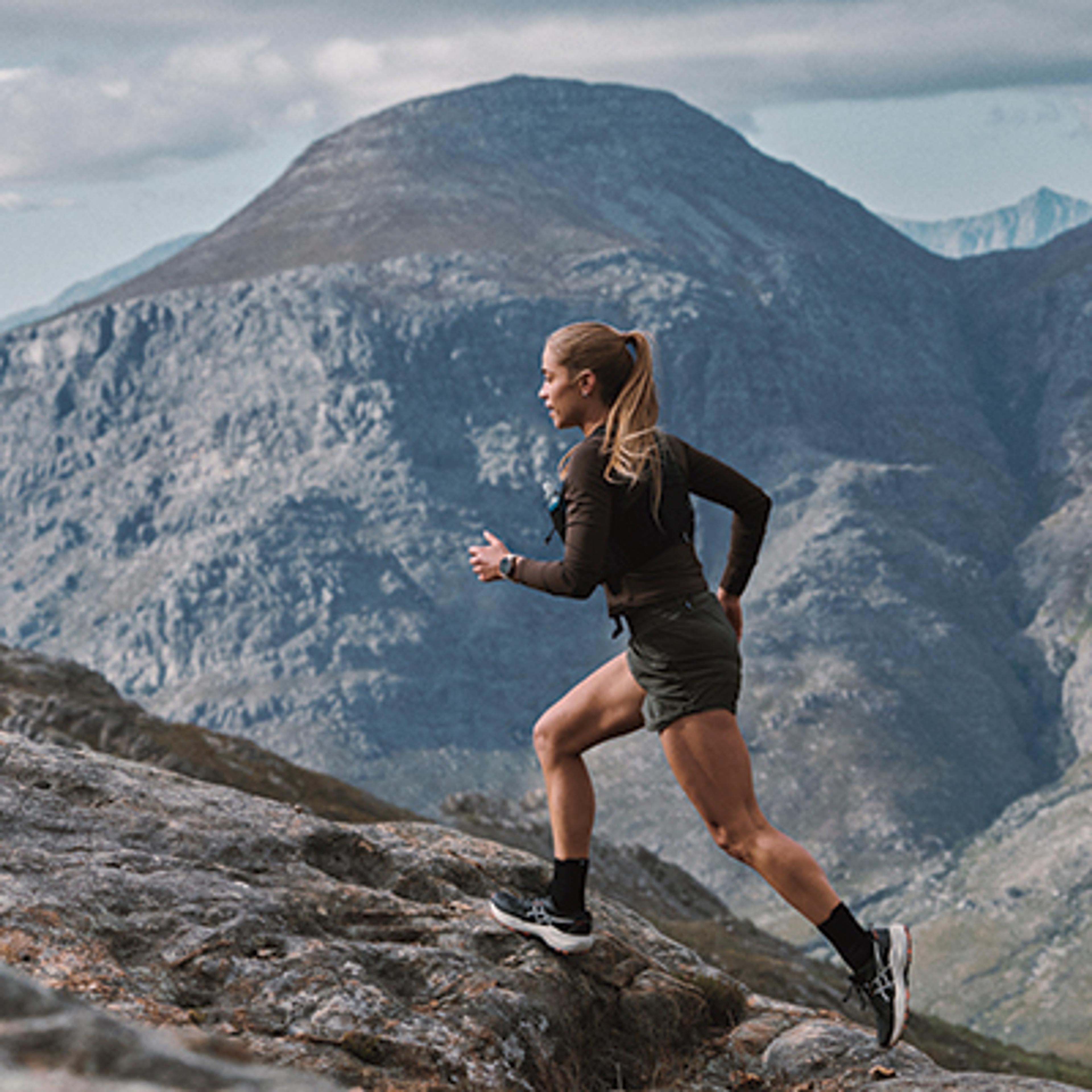 A woman trail running uphill on a rocky path with mountains in the background.