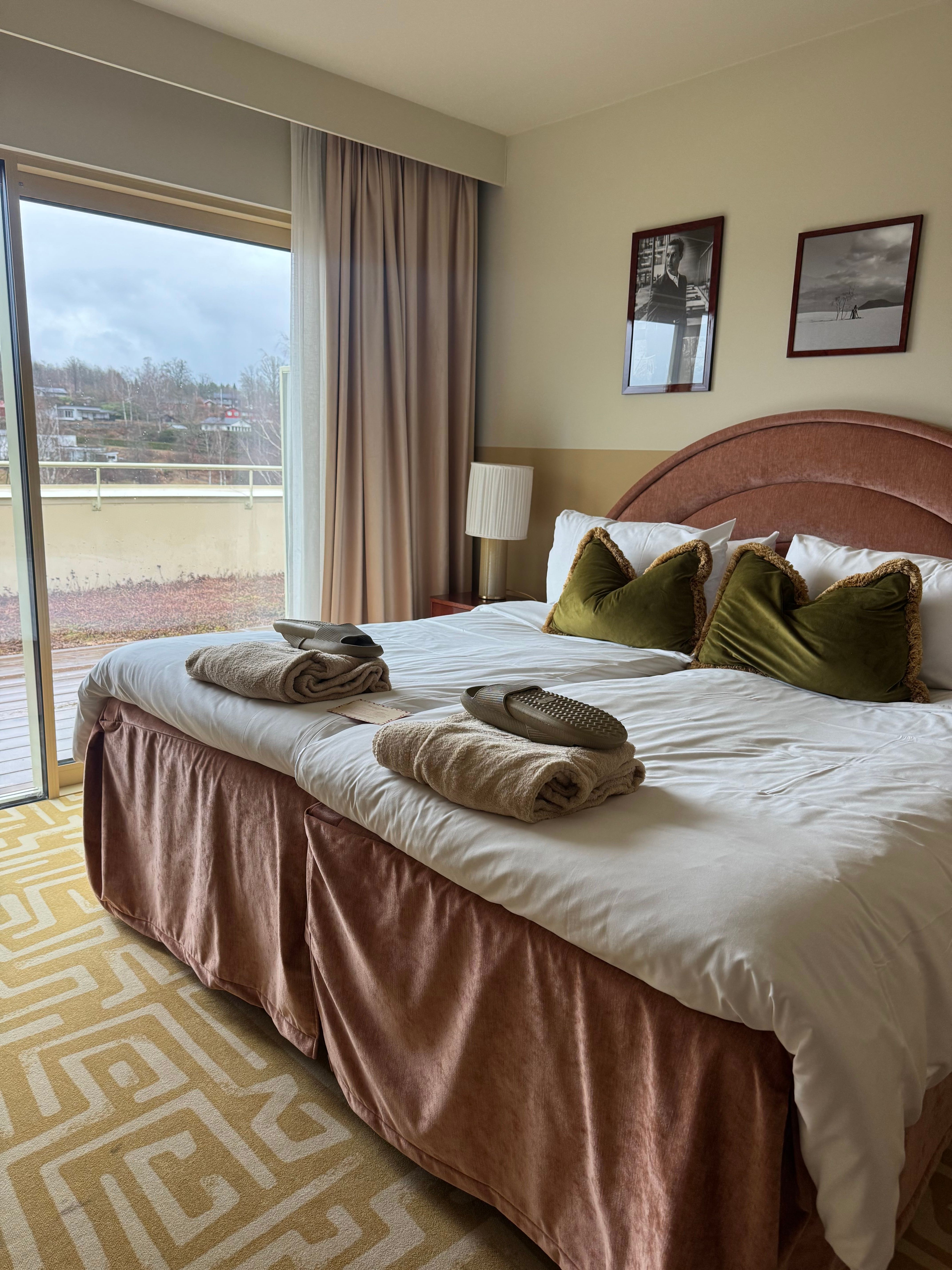 A hotel bedroom with a large bed featuring a pink headboard, white bedding, green pillows, towels, and slippers. A sliding glass door opens to a balcony overlooking a green hillside, with a geometric yellow rug on the floor.