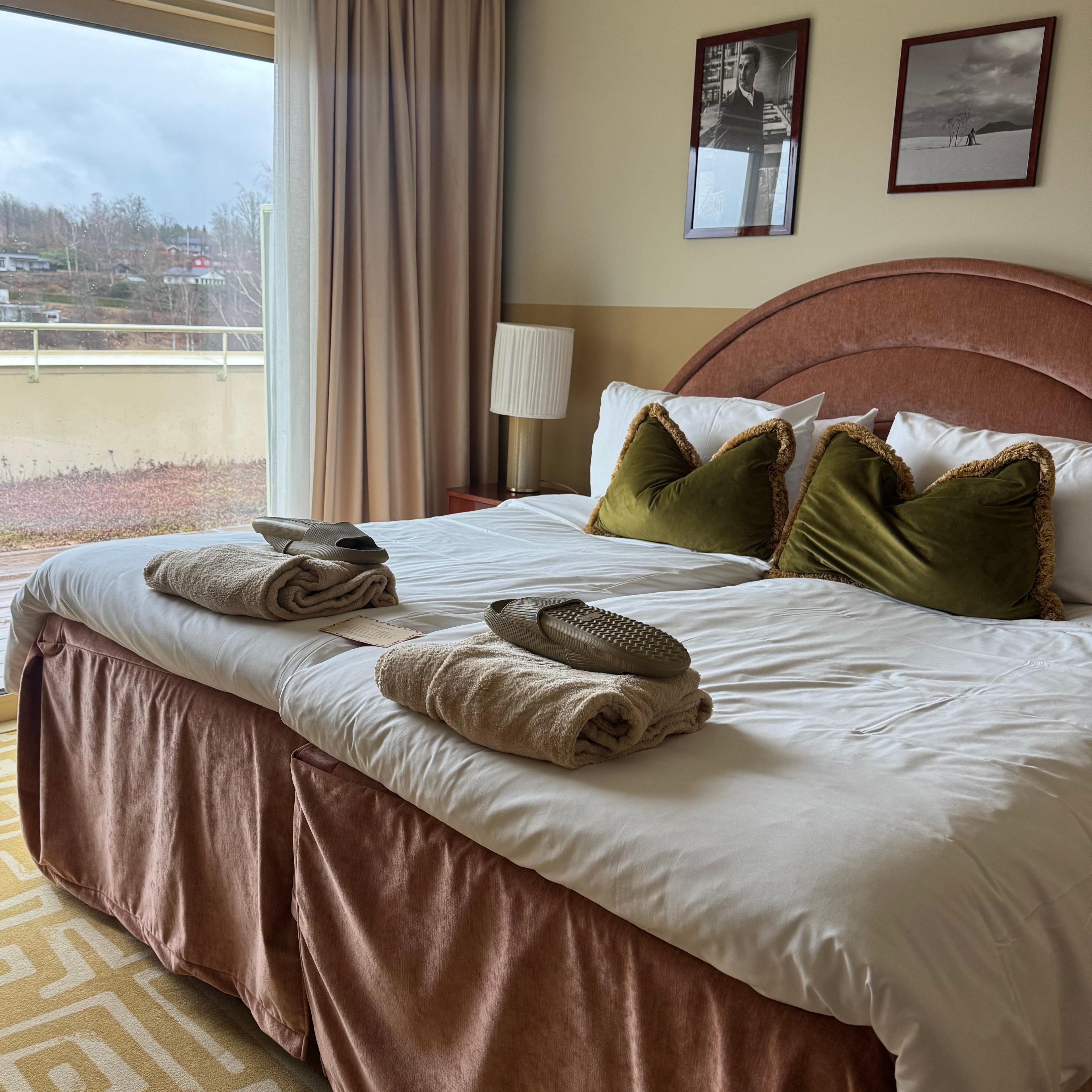 A hotel bedroom with a large bed featuring a pink headboard, white bedding, green pillows, towels, and slippers. A sliding glass door opens to a balcony overlooking a green hillside, with a geometric yellow rug on the floor.