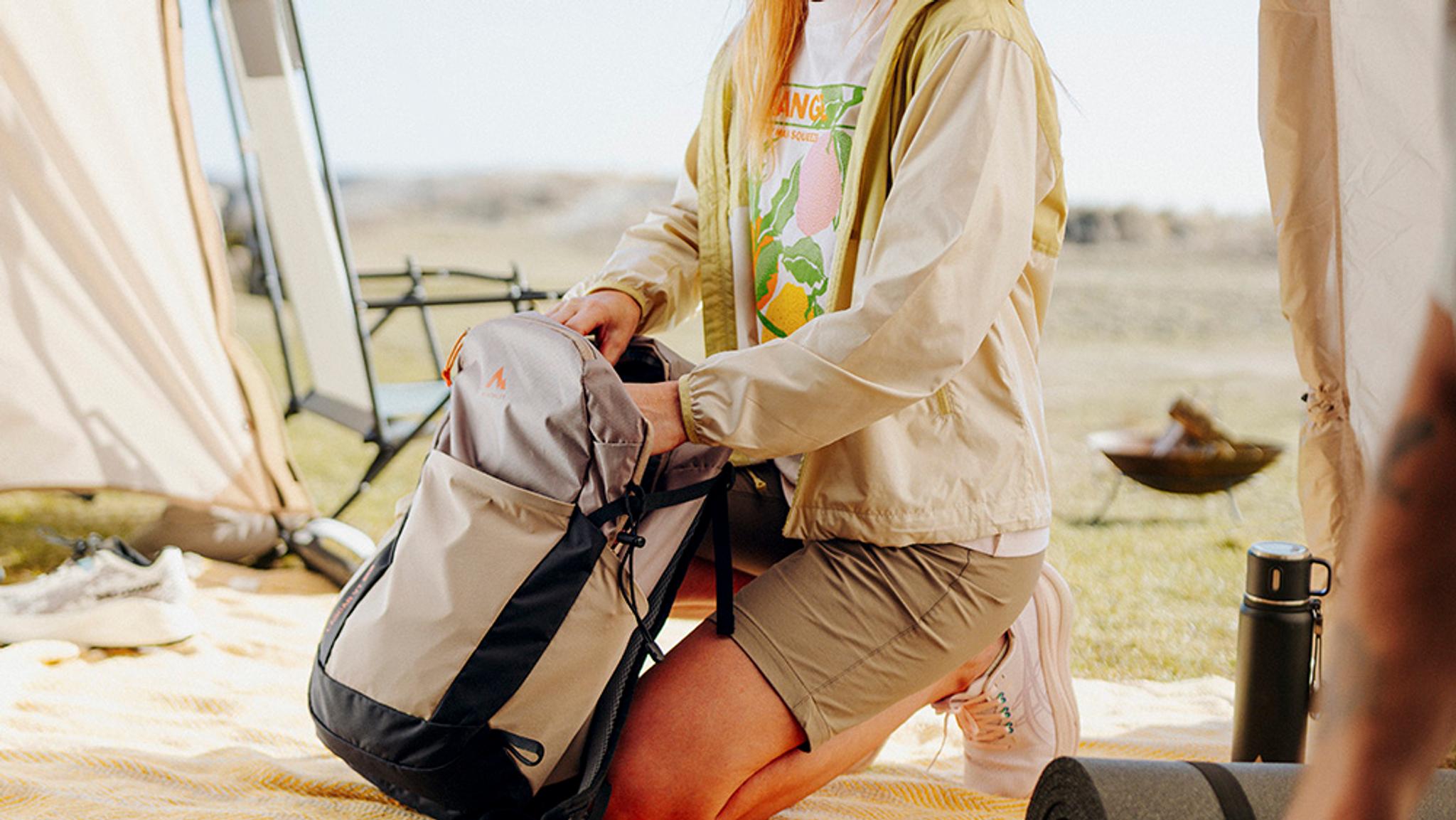 A person kneels, packing a tan and black backpack outdoors near a tent.