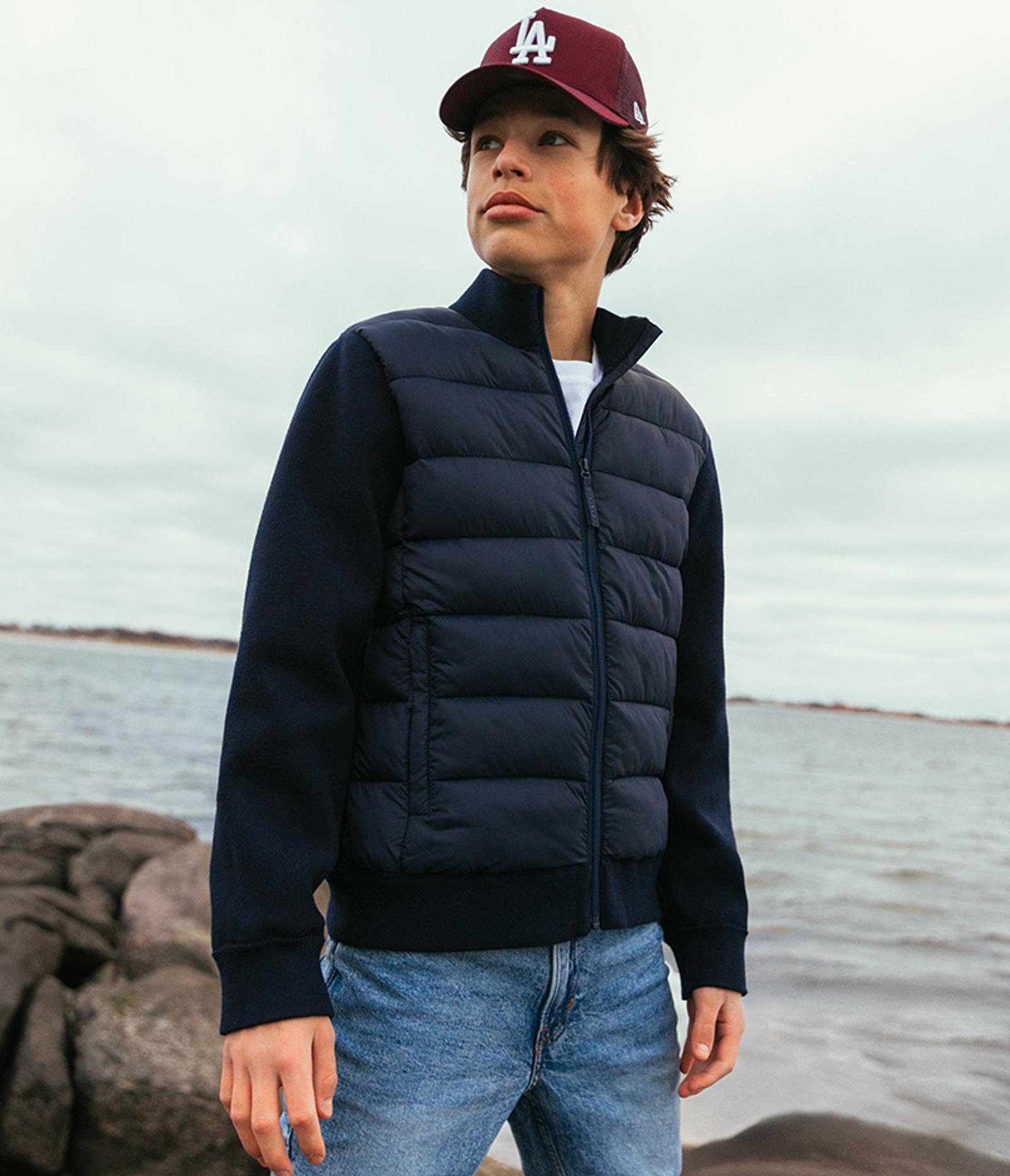 A young boy in a dark blue puffer jacket with knitted sleeves, blue jeans, and a maroon baseball cap stands on rocks by the water, looking left.