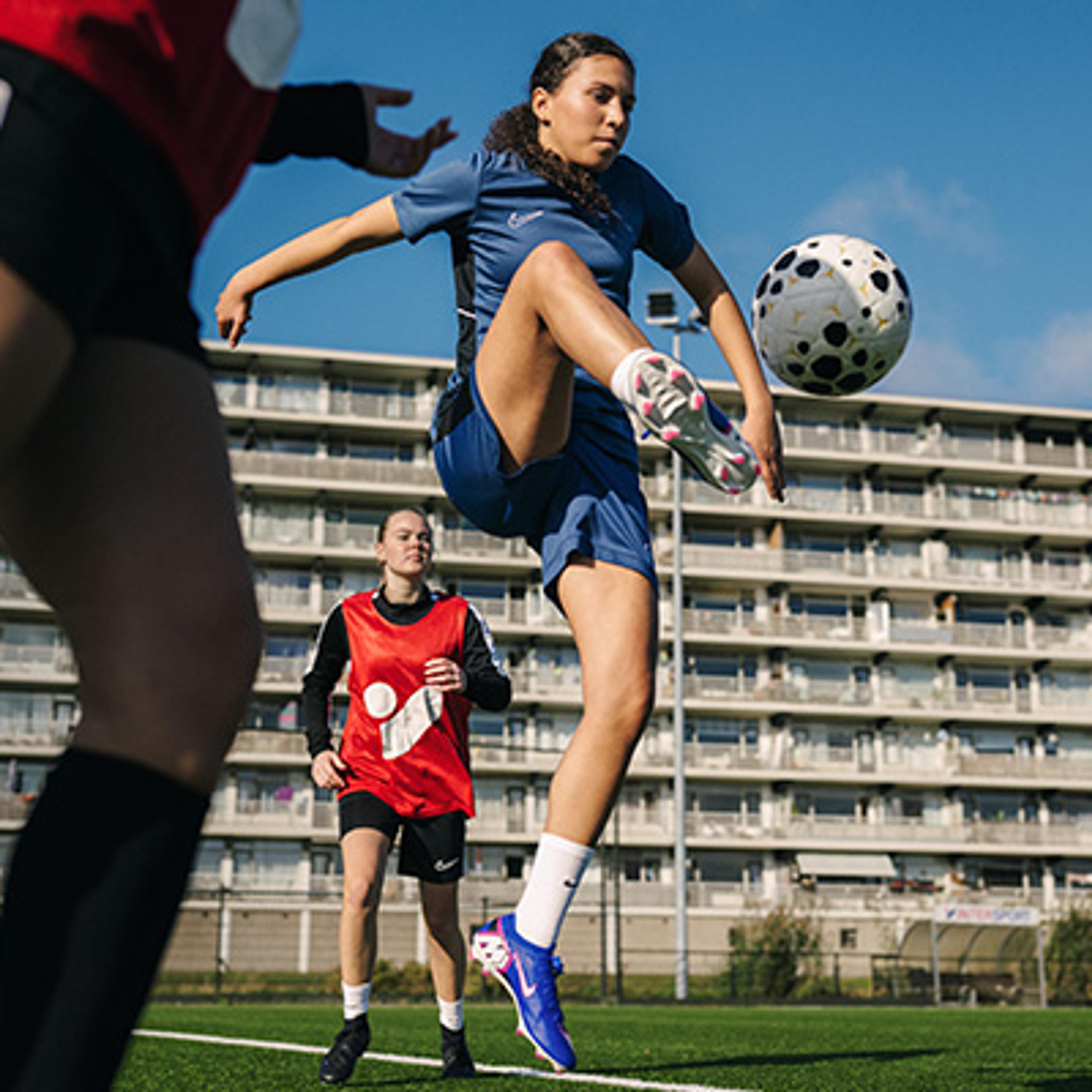 A young woman in a blue uniform kicks a soccer ball mid-air on a turf field, with other players and an apartment building in the background.