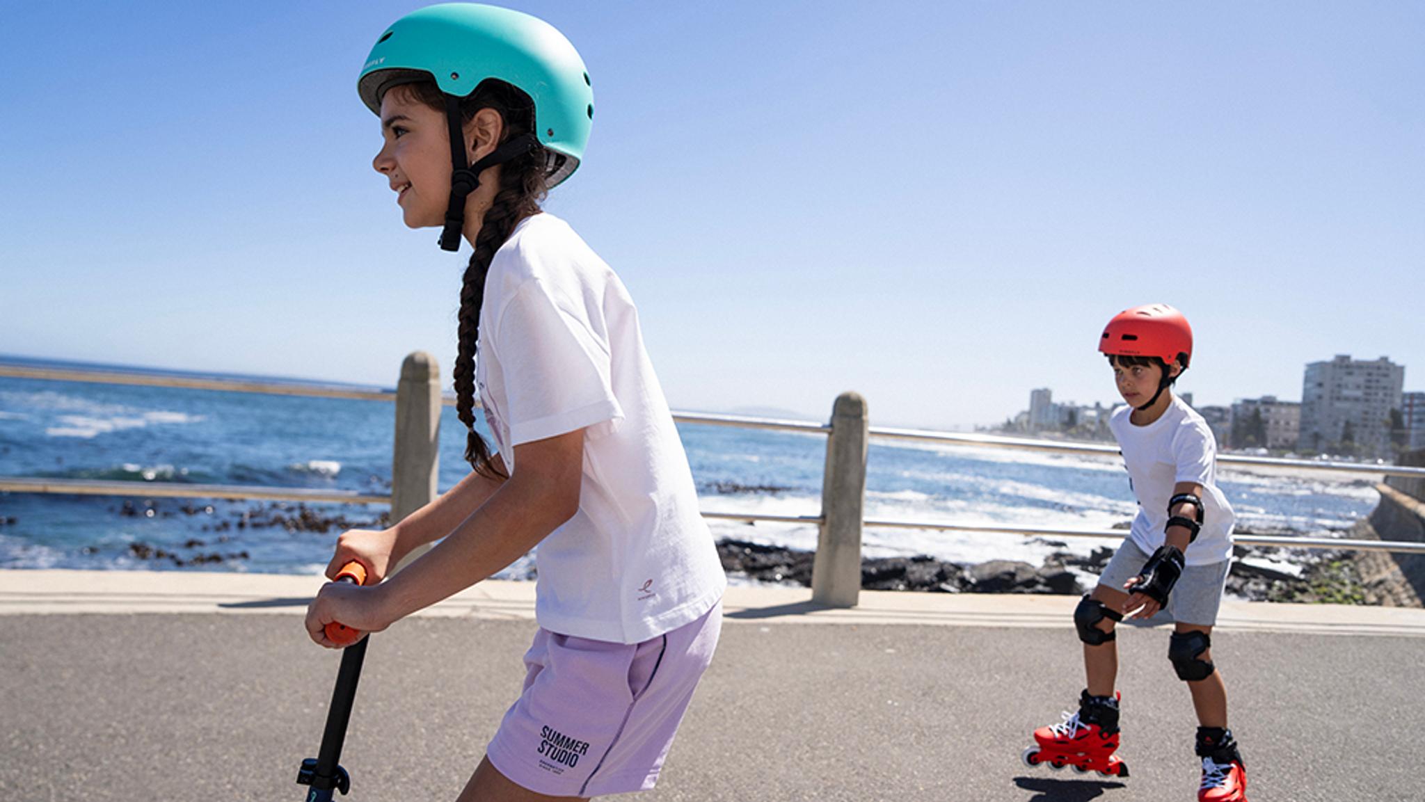 Girl on a scooter and boy rollerblading on a sunny seaside promenade.
