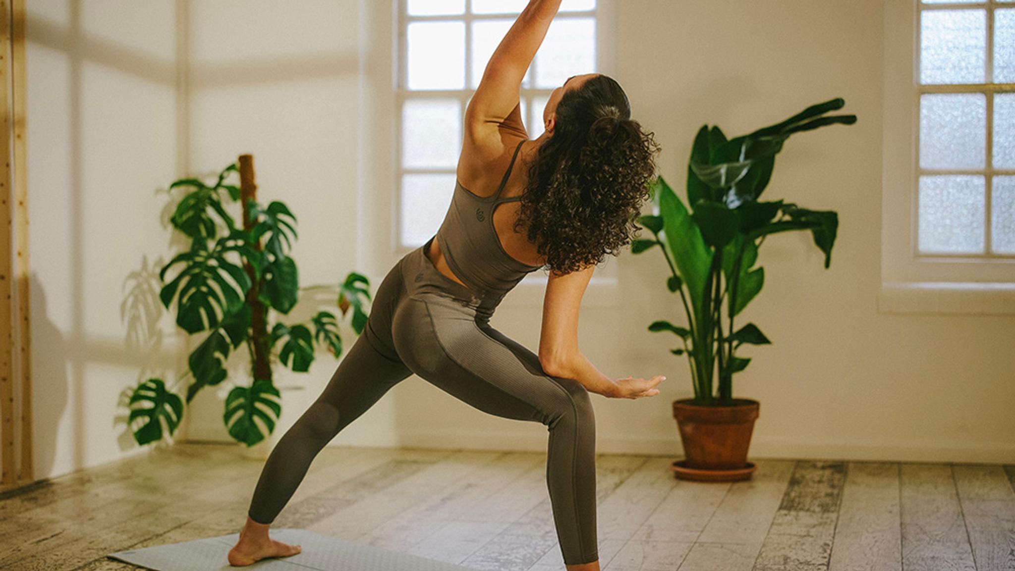 A woman in grey activewear performing a yoga pose with one arm raised in a room with plants.