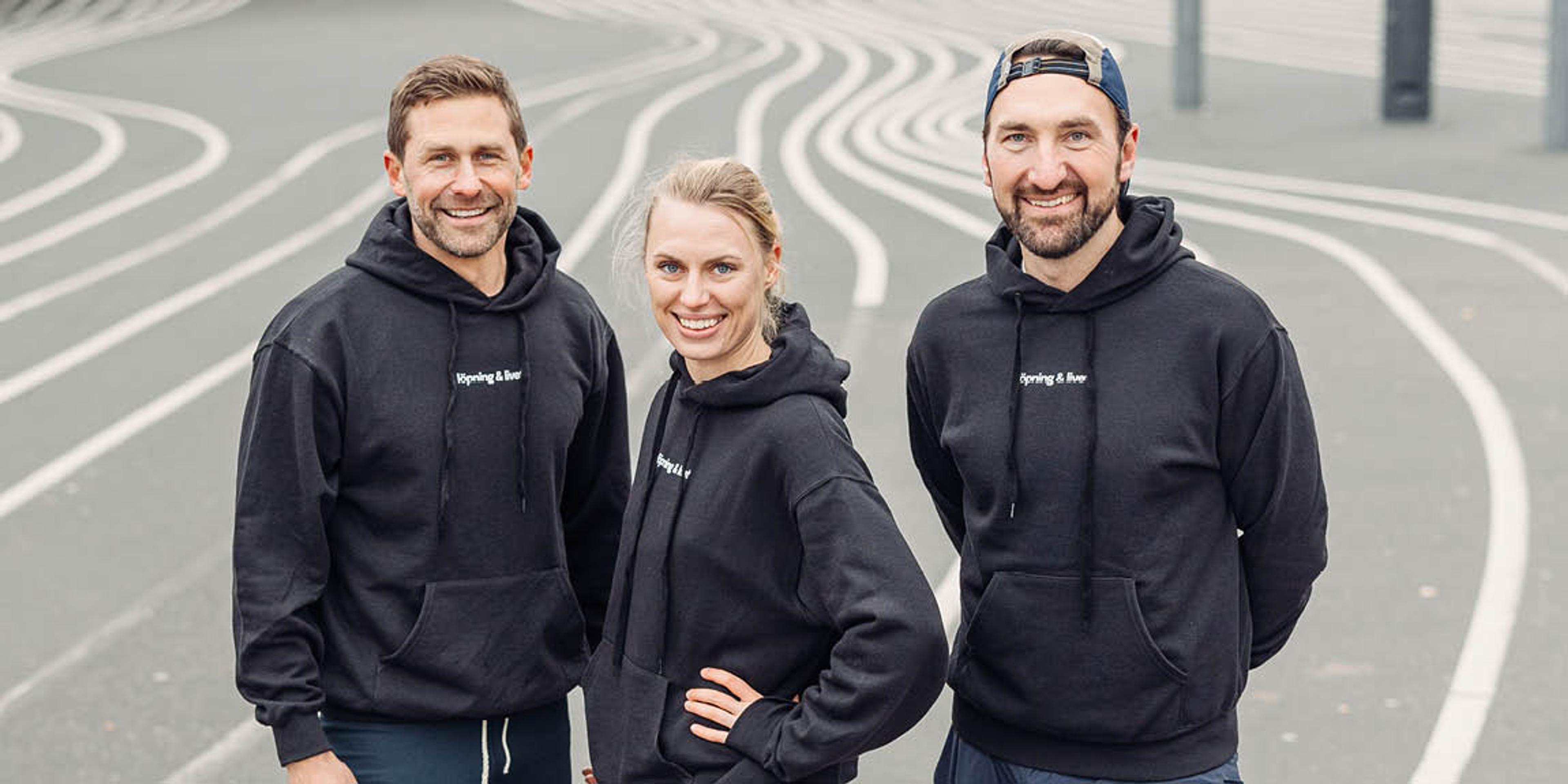 Two men and one woman smiling in matching black hoodies on an outdoor surface with wavy white lines.