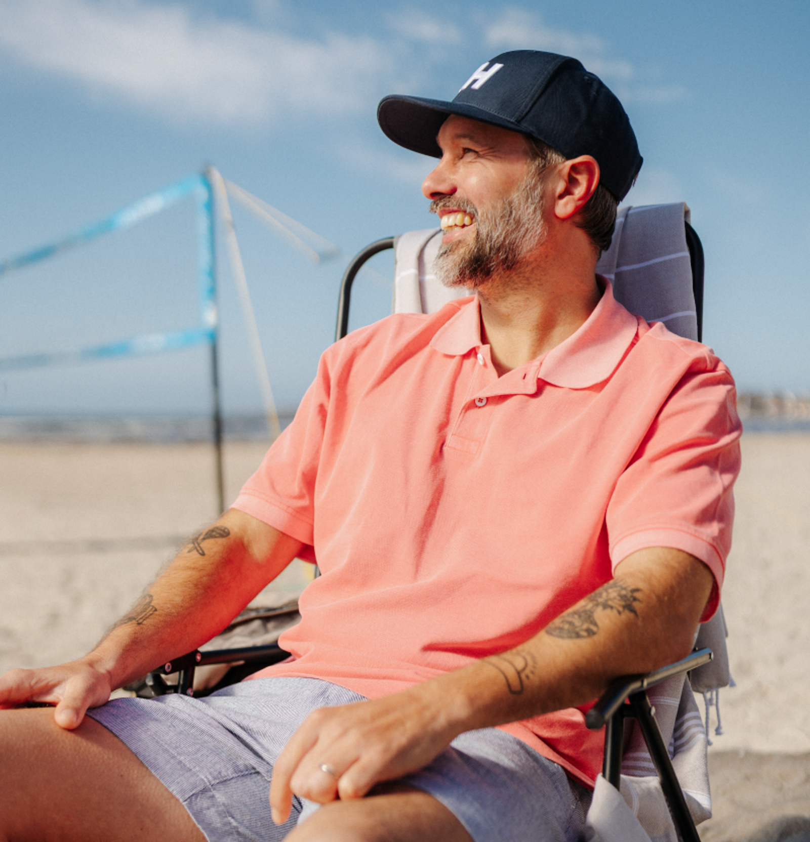 Smiling man in a navy H-logo cap and peach polo relaxing on a beach.