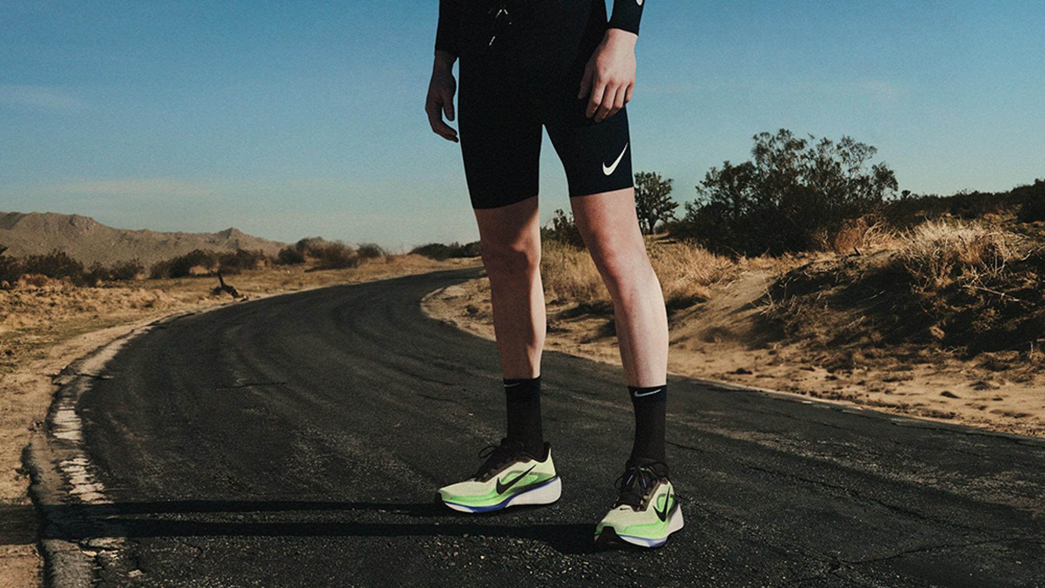 A person in black shorts and bright green Nike running shoes stands on a curved desert road.