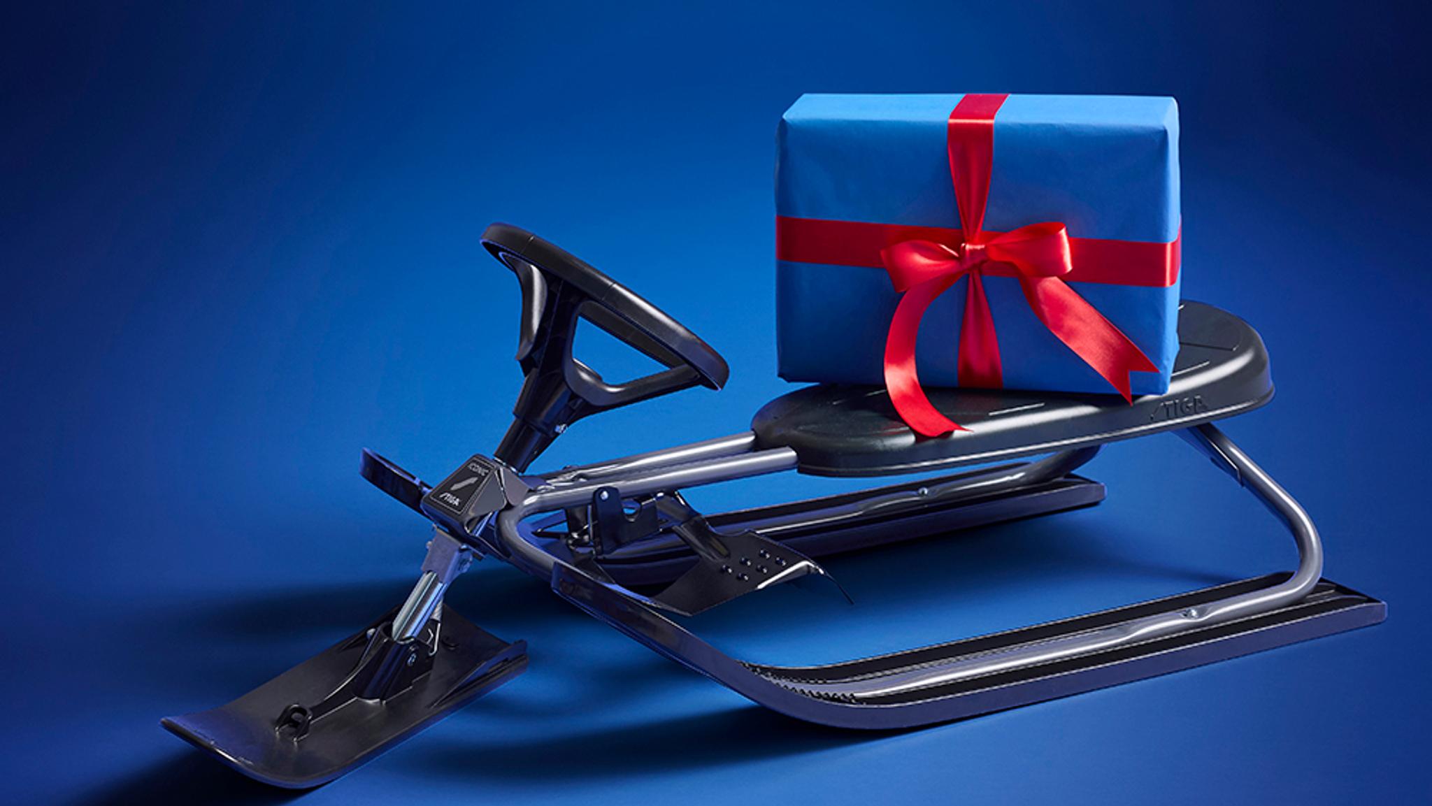 A blue gift box with a red ribbon rests on a dark grey steerable snow sled.