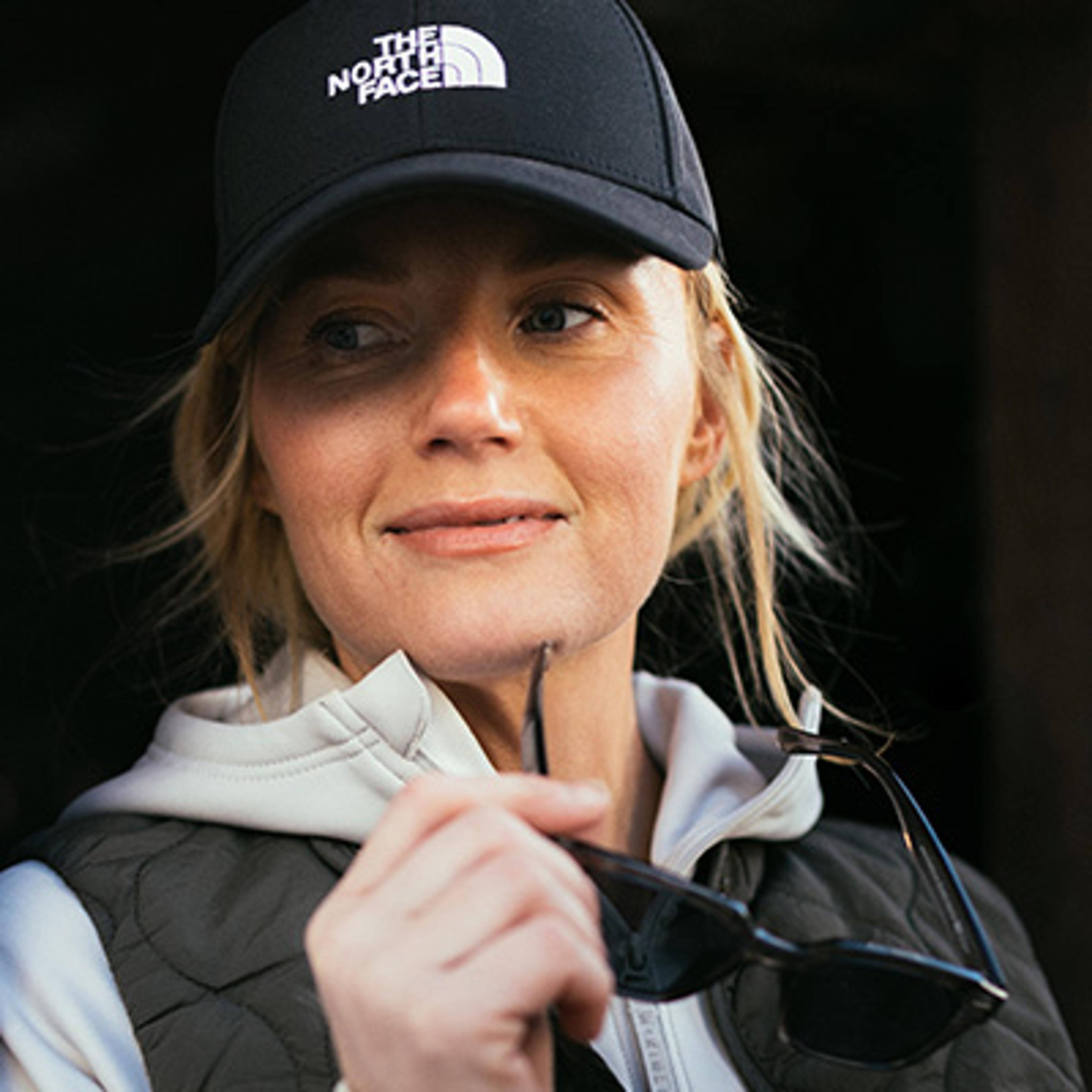 A woman in a black The North Face cap holds sunglasses, looking slightly left with a subtle smile.