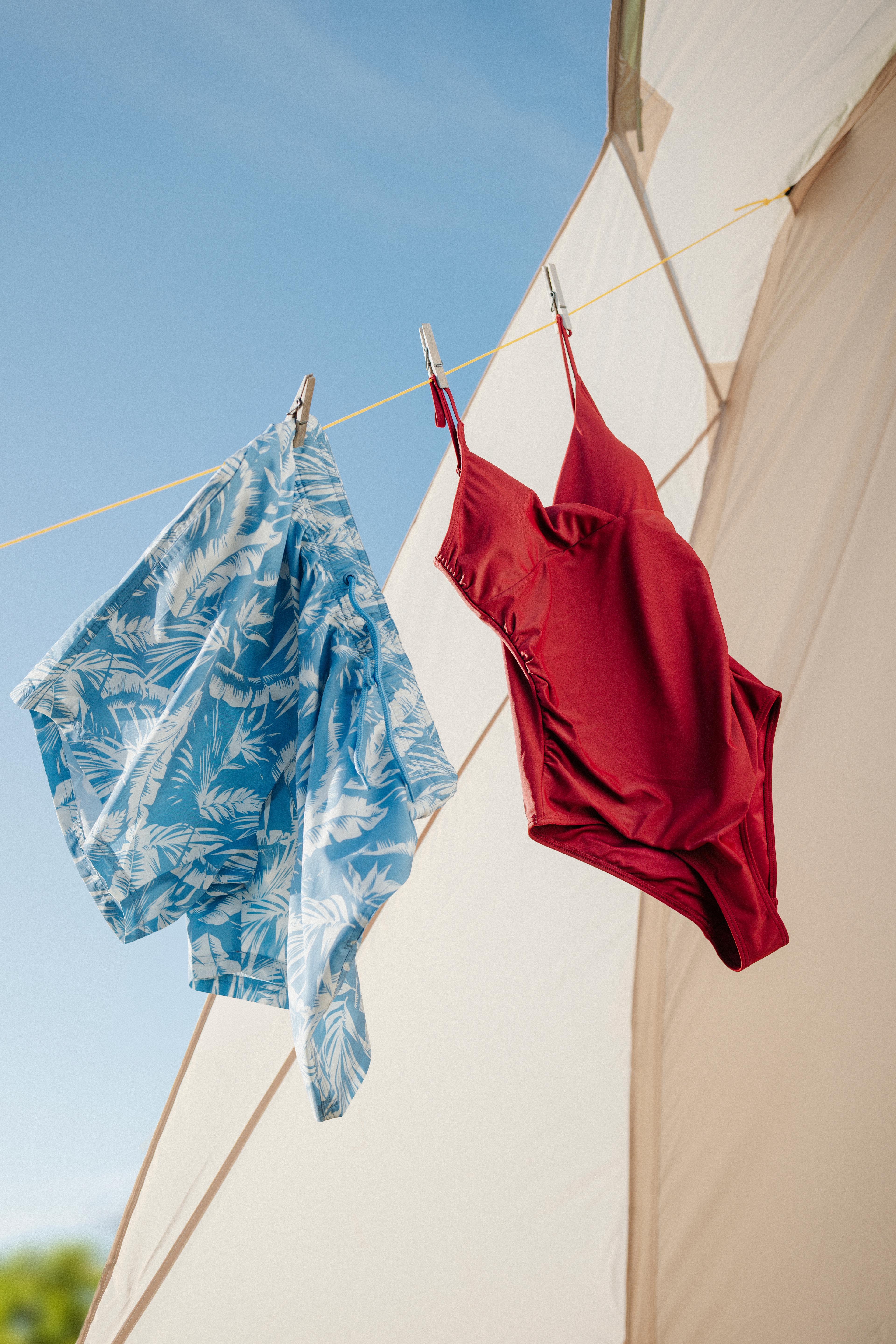 A blue tropical print shirt and a red swimsuit hang on a clothesline against a blue sky and beige tent.