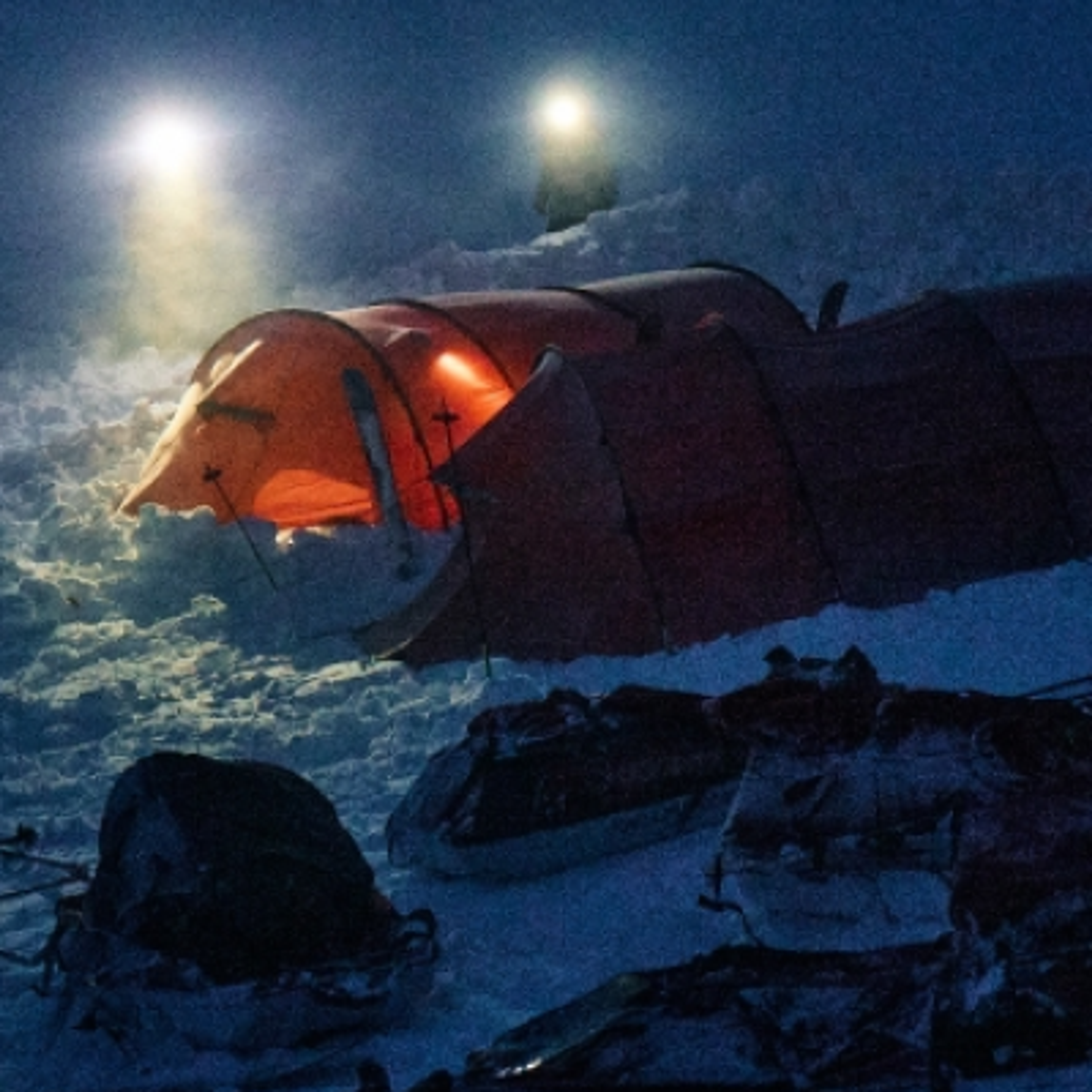 Glowing orange tent in a snowy night camp with distant headlamps.
