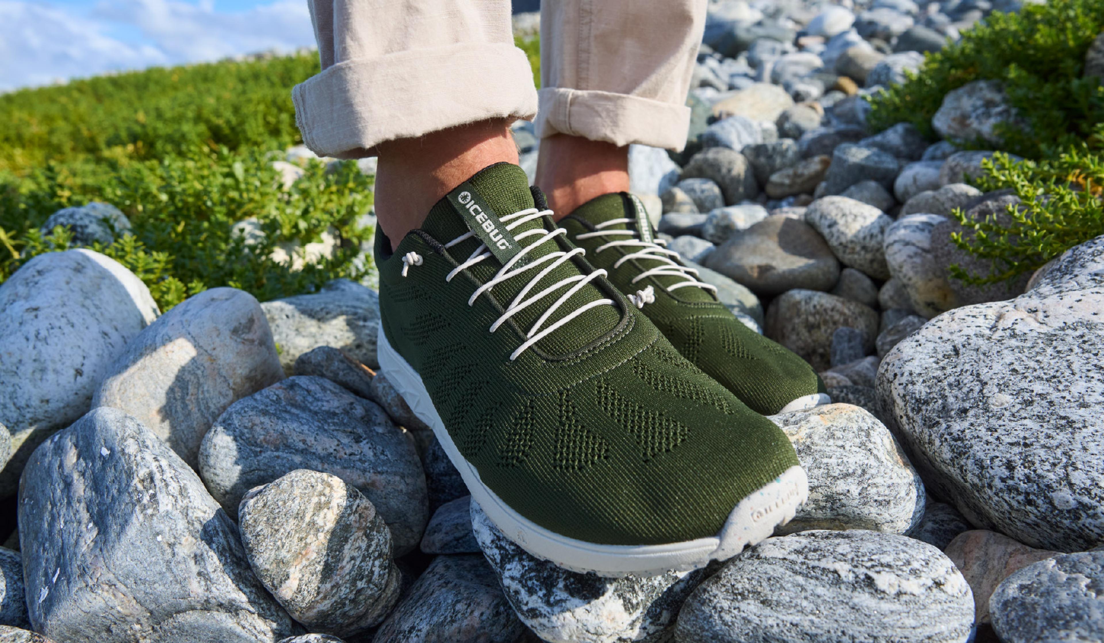 Person wearing green ICEBUG shoes with white laces, standing on rocks.