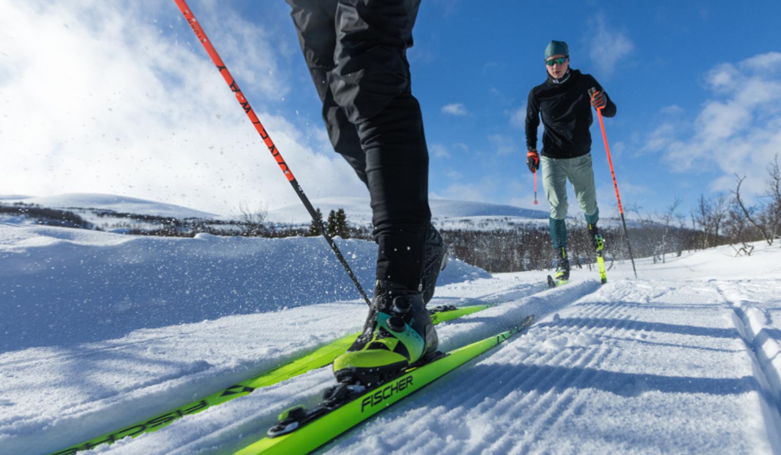 Two people cross-country skiing on a sunny, snowy path, with bright yellow skis in the foreground kicking up snow.