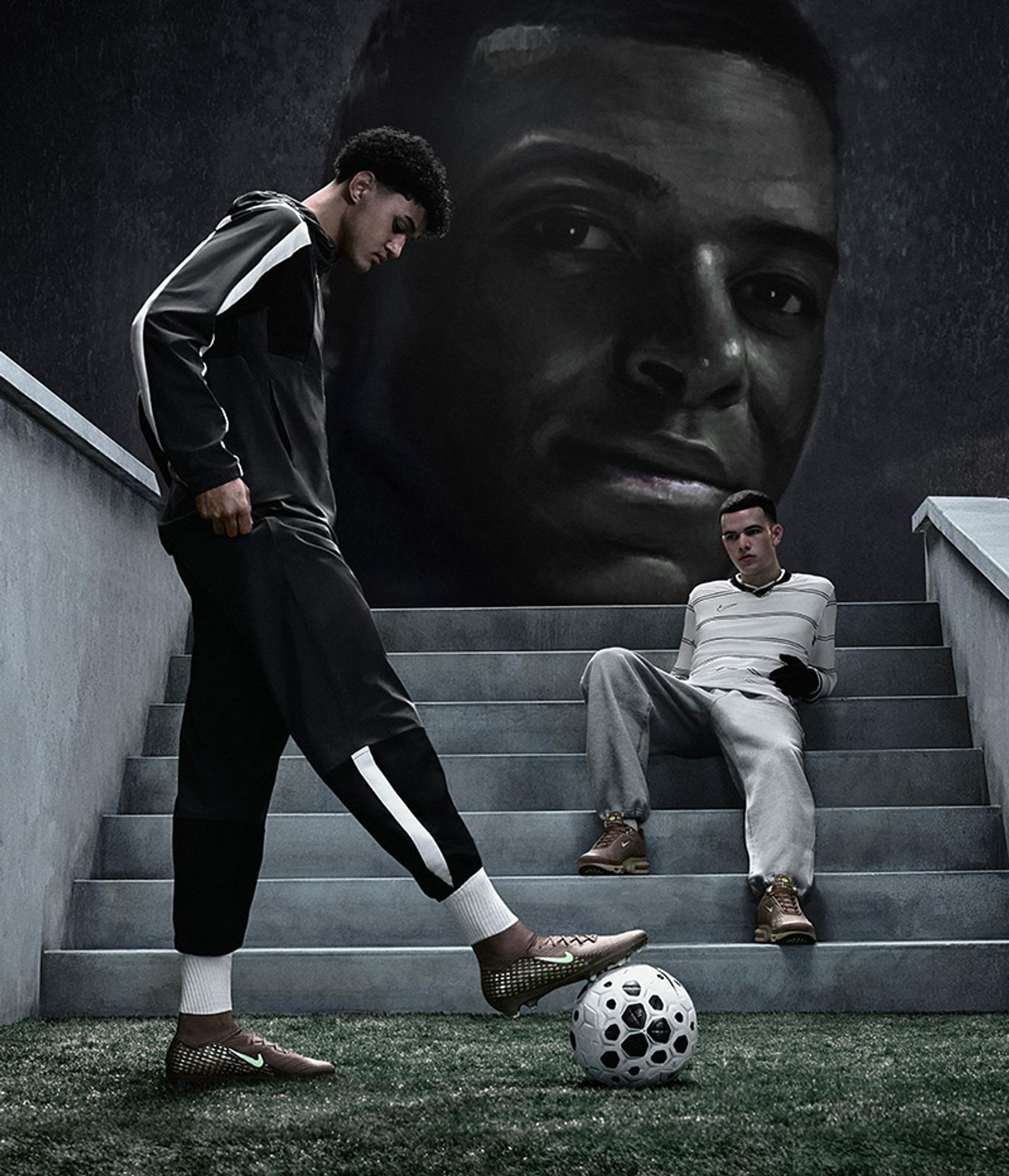 A young man stands with his foot on a soccer ball on artificial grass, next to stairs where another man sits, all beneath a large dark mural of a man's face.