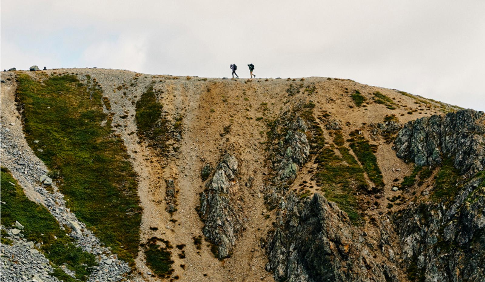 Two hikers walk with backpacks along a rocky mountain ridge.