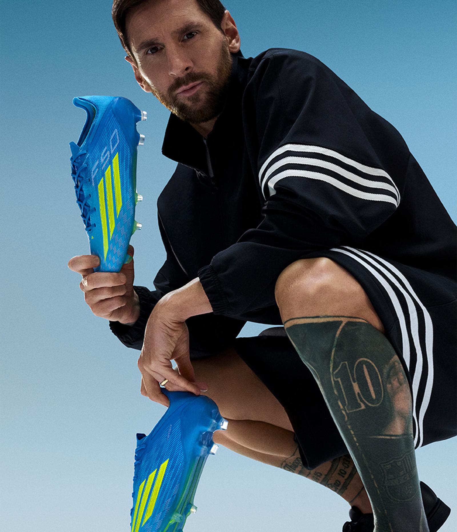 Lionel Messi in black Adidas sportswear, crouching and holding blue and yellow soccer cleats.