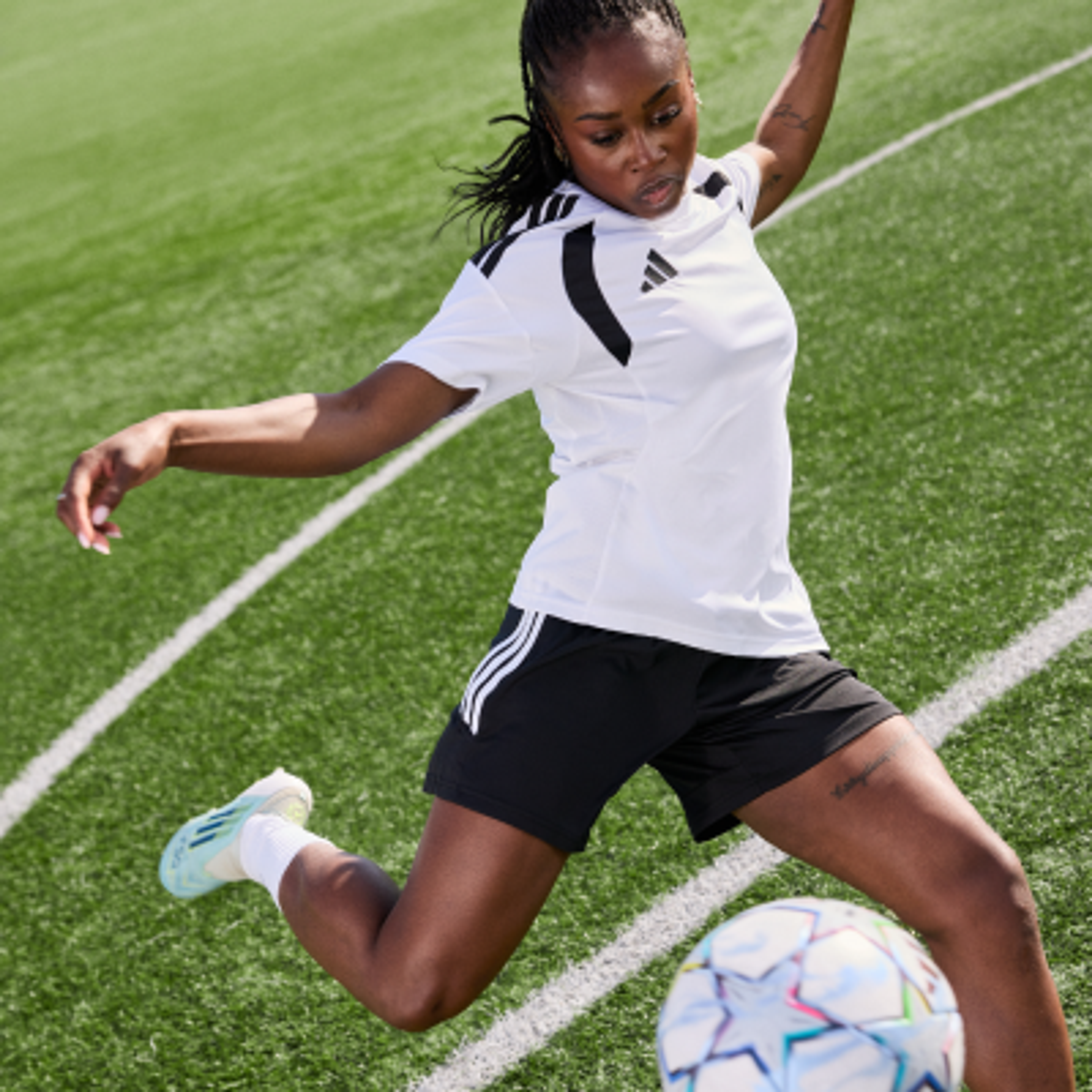 A woman in a white Adidas shirt and black shorts kicks a soccer ball on a green field.