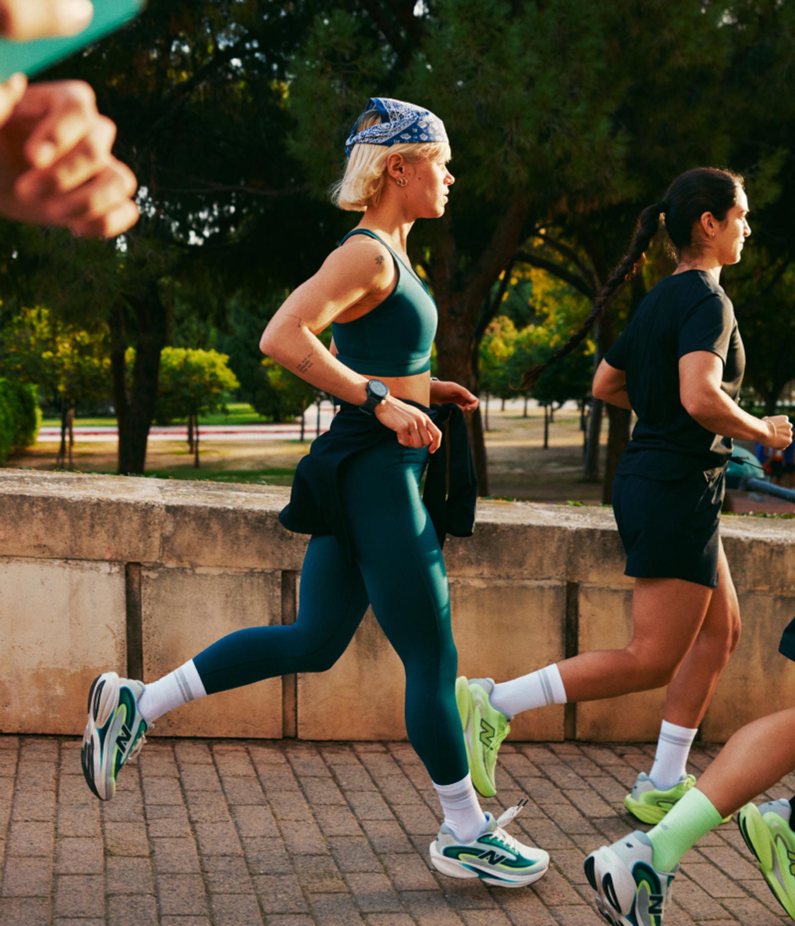 Three women are running outdoors on a paved path next to a stone wall and trees.