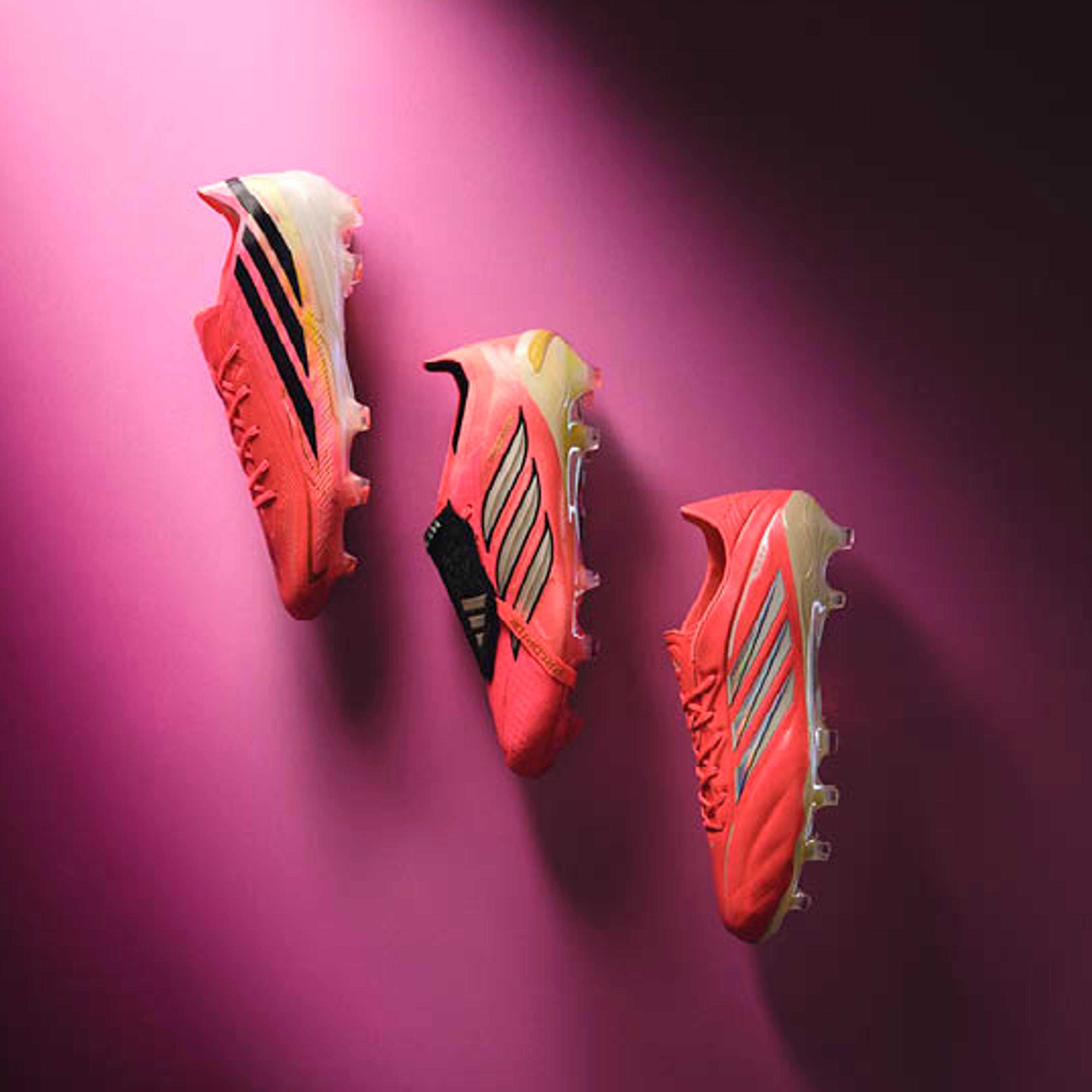 Three coral Adidas football boots arranged diagonally on a pink and purple gradient background.