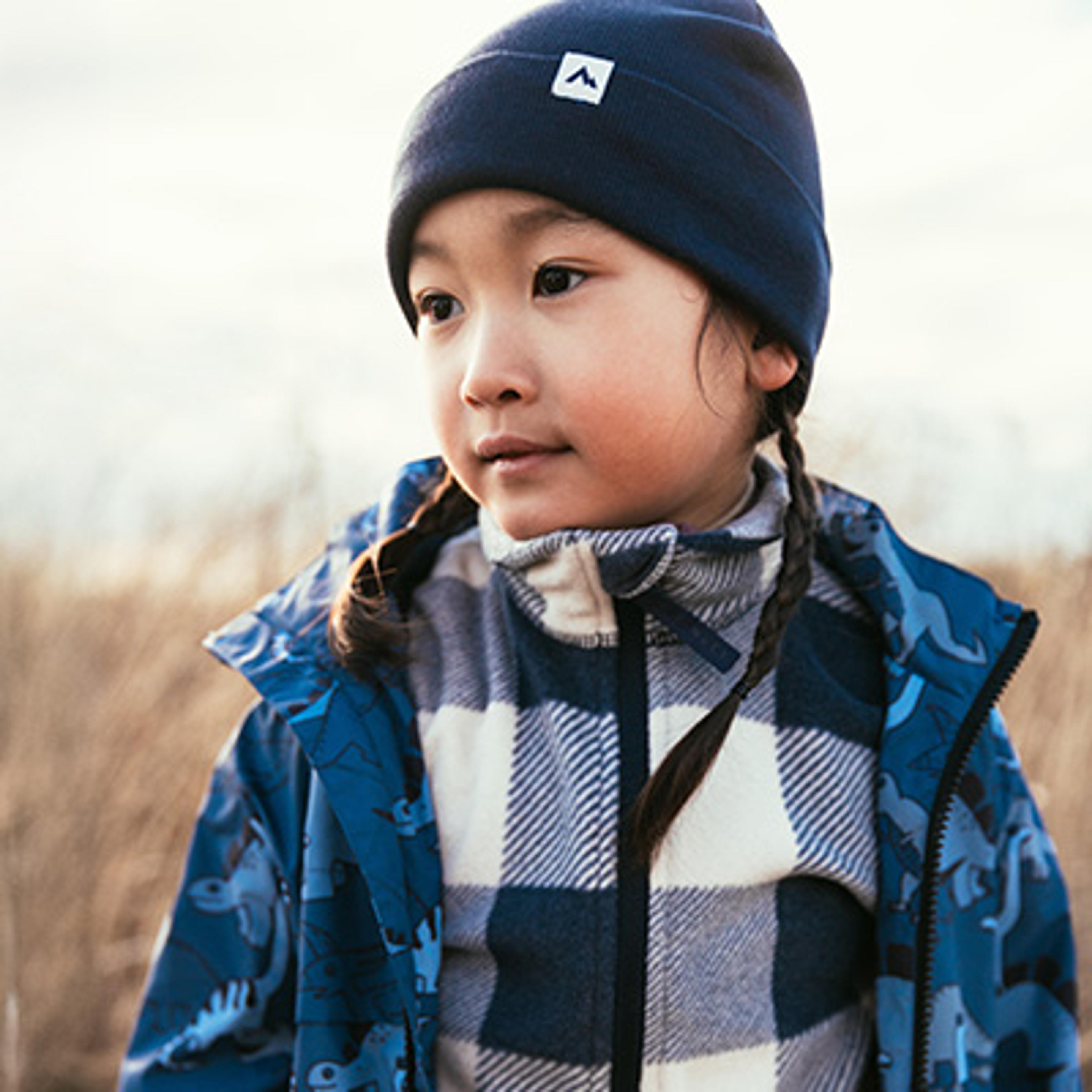 A child in a blue beanie, plaid fleece, and dinosaur jacket looks right outdoors.