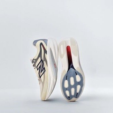 Two New Balance sneakers: one upright, displaying a beige, gray, and blue wavy upper, and the other revealing its cream sole with blue traction and a red detail.