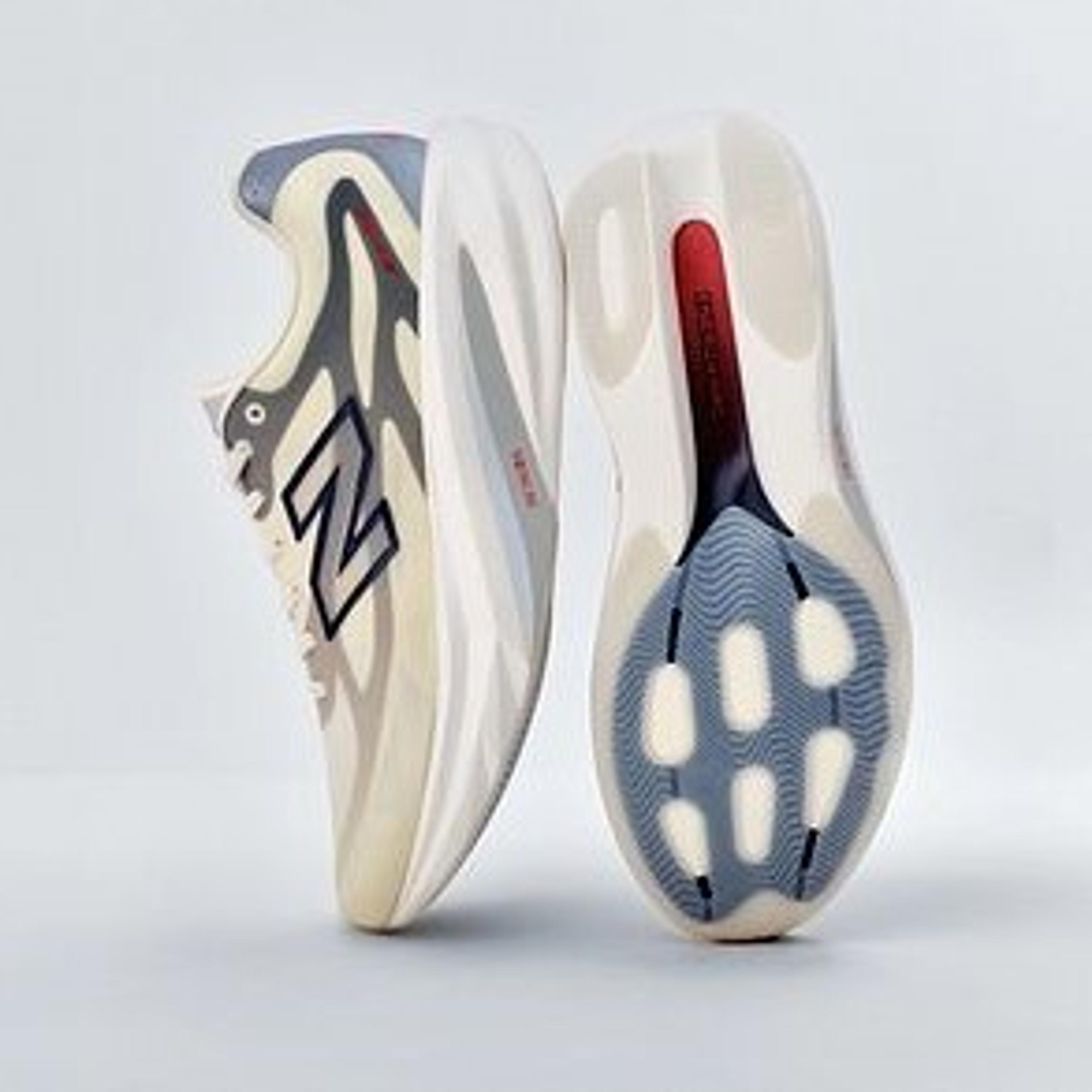 Two New Balance sneakers: one upright, displaying a beige, gray, and blue wavy upper, and the other revealing its cream sole with blue traction and a red detail.