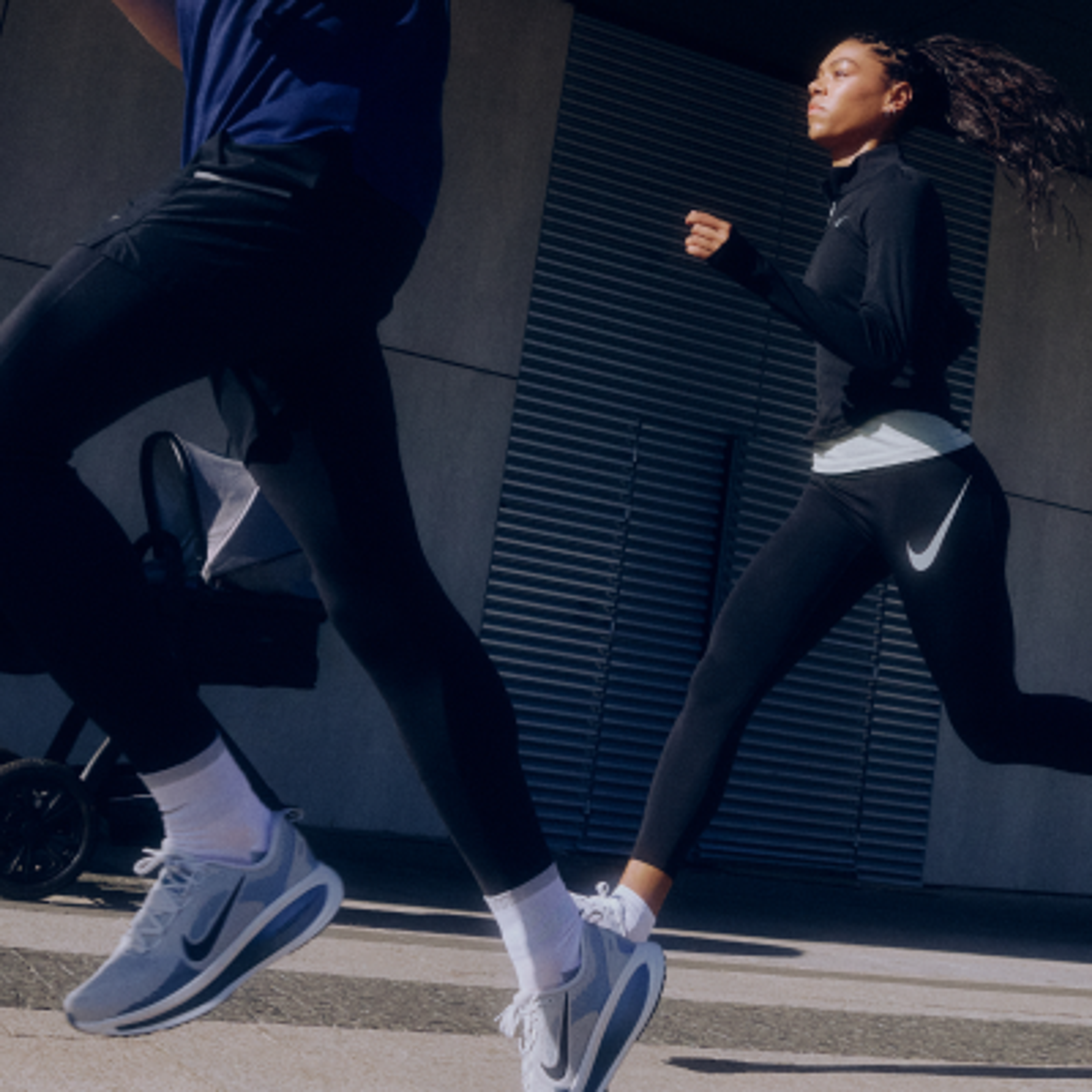 A woman runs alongside another person pushing a stroller, both wearing Nike athletic wear outdoors.