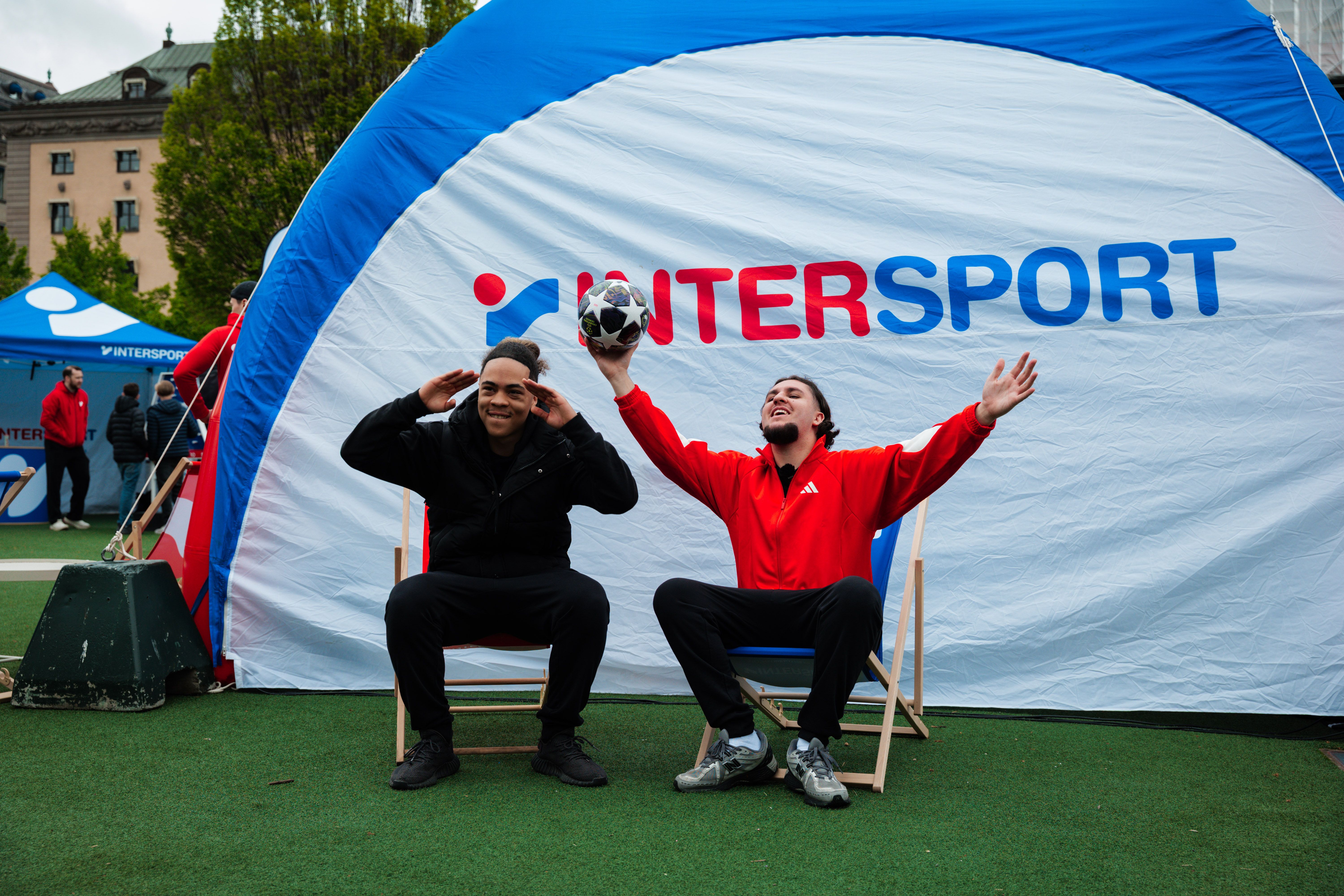 Two joyful men pose in front of an Intersport tent, one holding a soccer ball aloft.