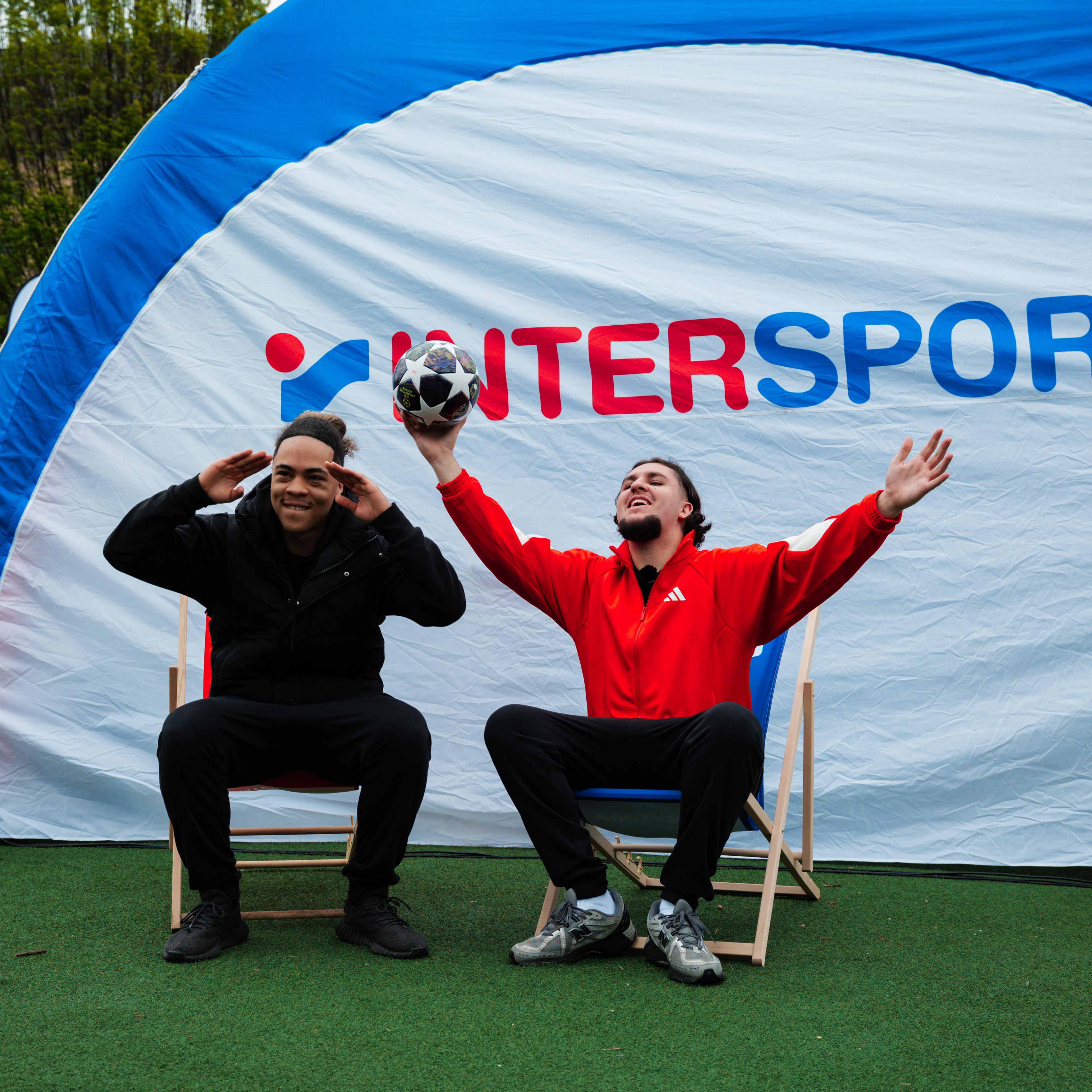 Two joyful men pose in front of an Intersport tent, one holding a soccer ball aloft.