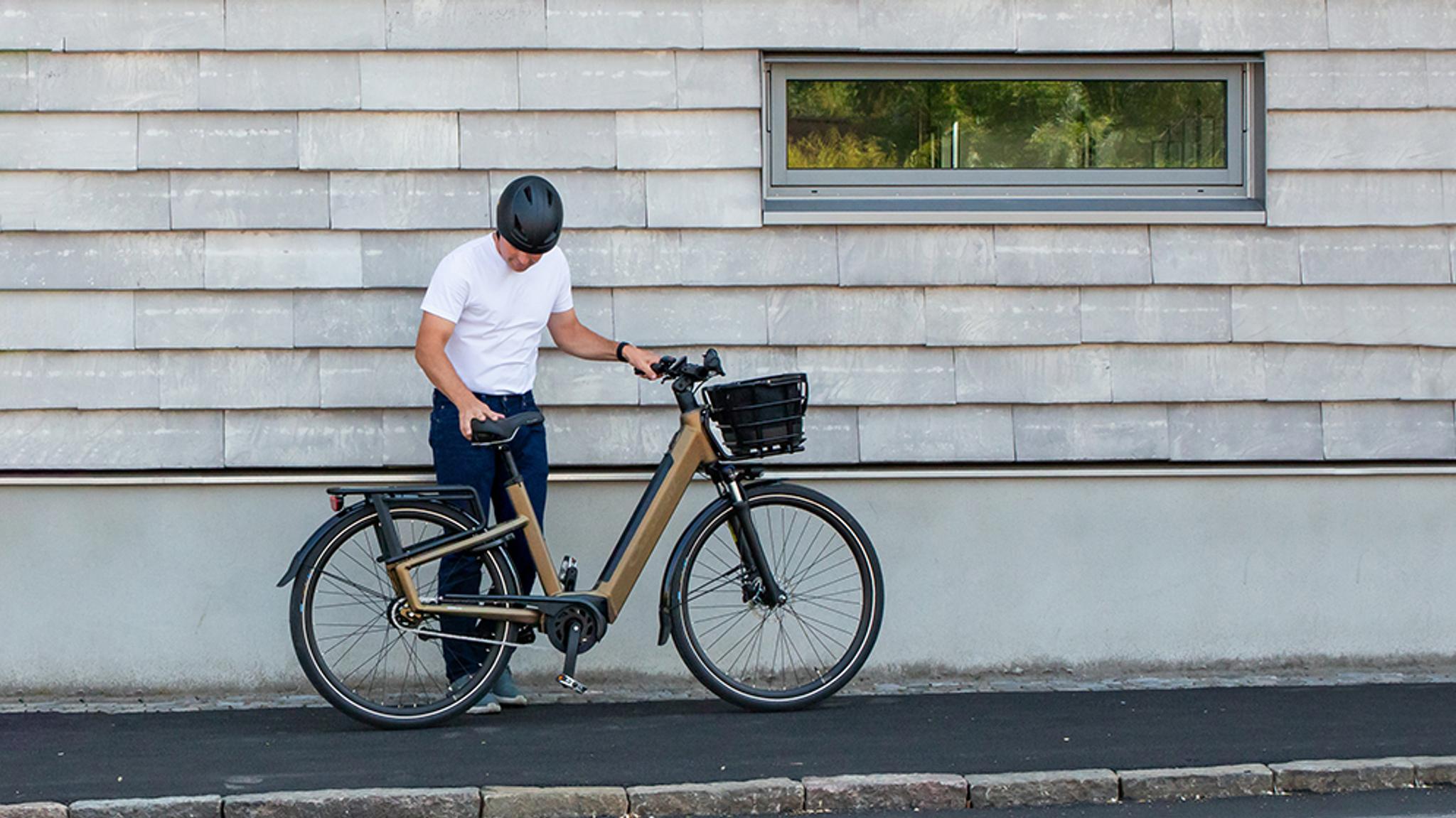 A man in a helmet and white shirt adjusts a gold electric bicycle with a front basket and rear rack on a sidewalk.