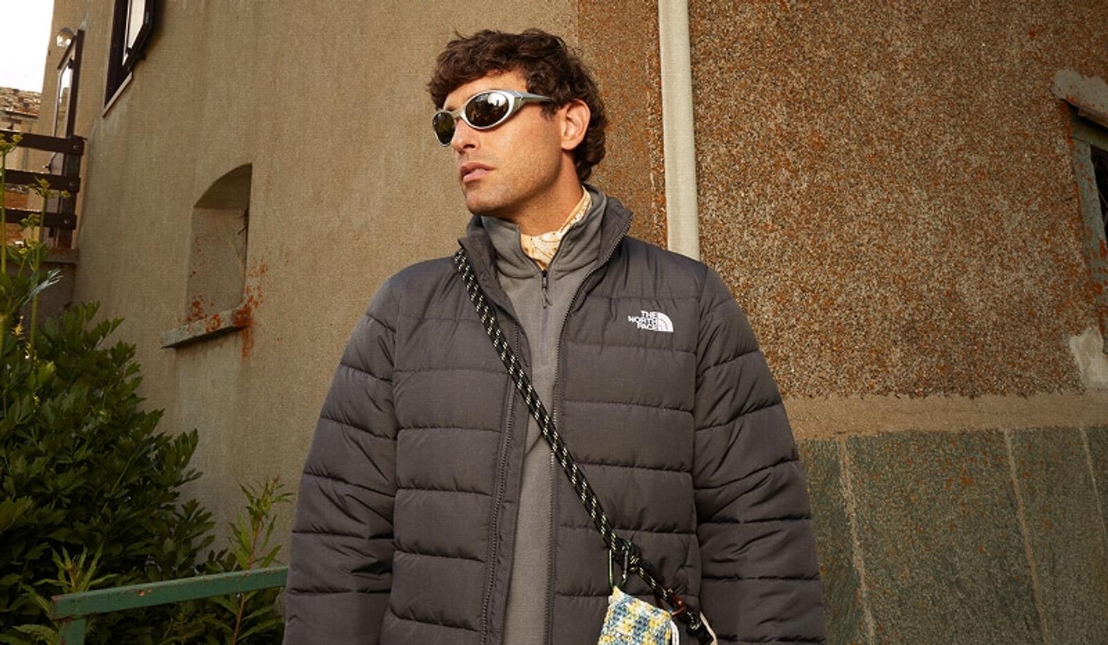 Man in a dark gray puffer jacket, sporty silver sunglasses, and curly brown hair, looking right against a background of old buildings.