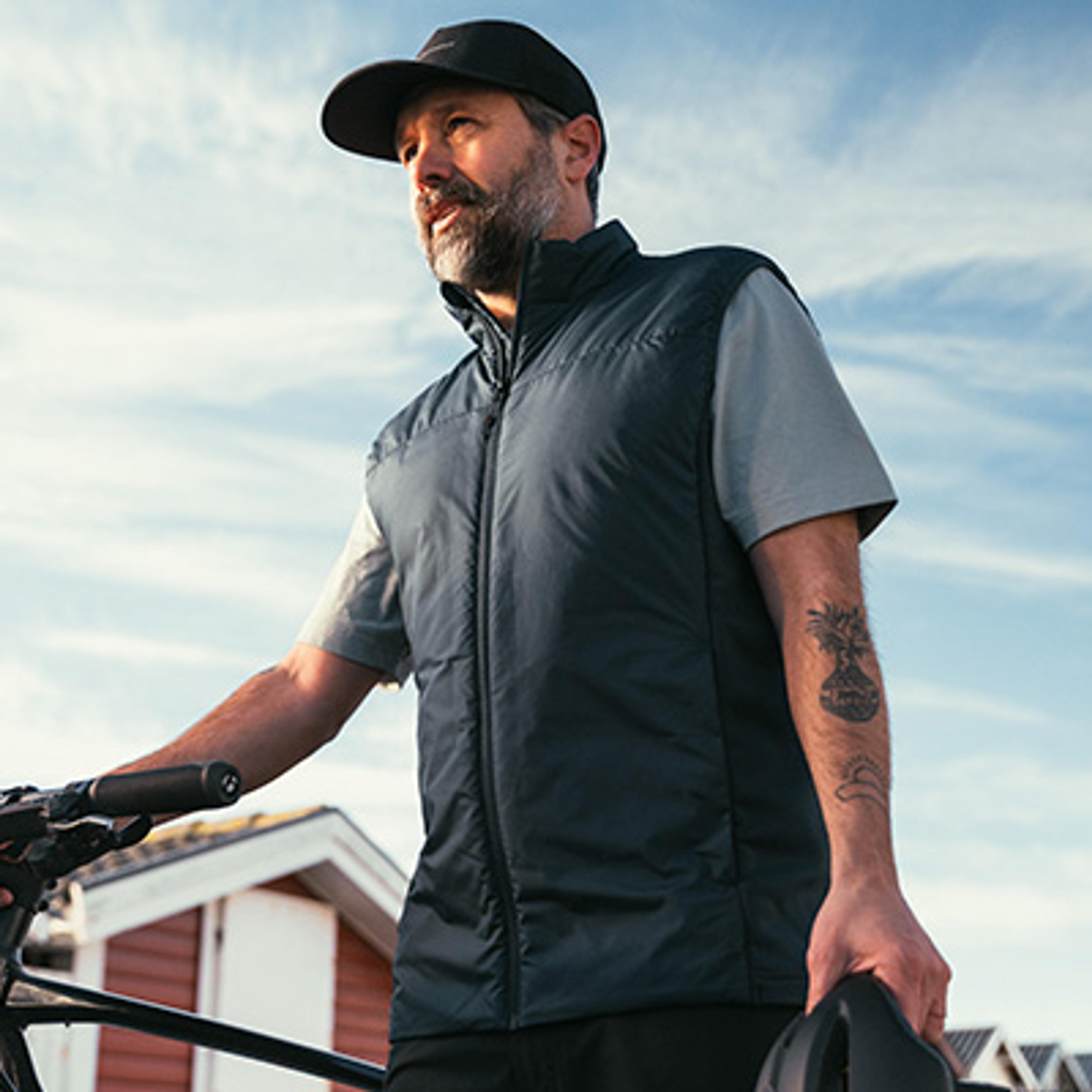 A bearded man in a dark vest and cap holds a bicycle and helmet outdoors.