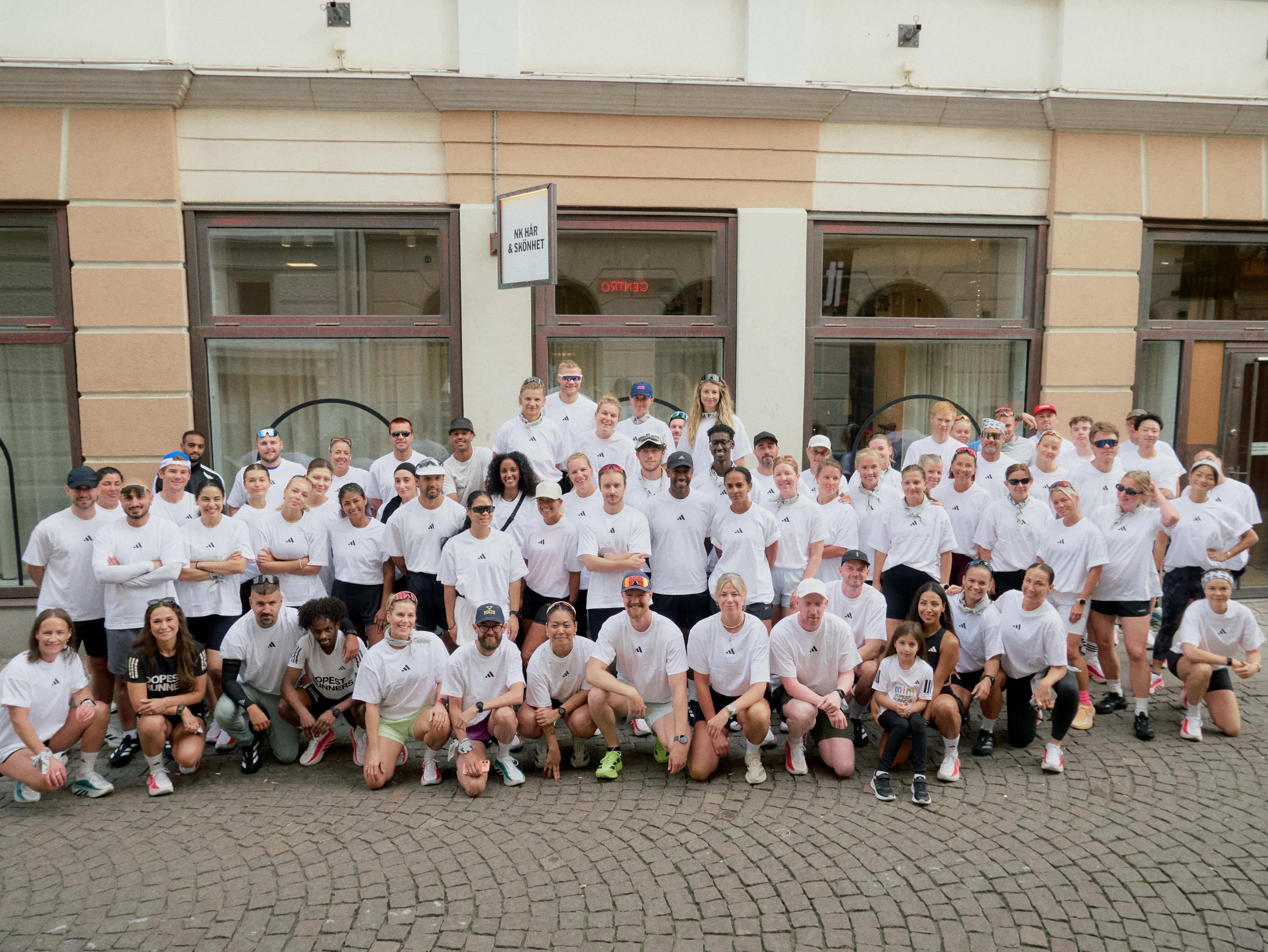 A large, diverse group of people, mostly wearing white t-shirts, pose together on a cobbled street in front of a building.