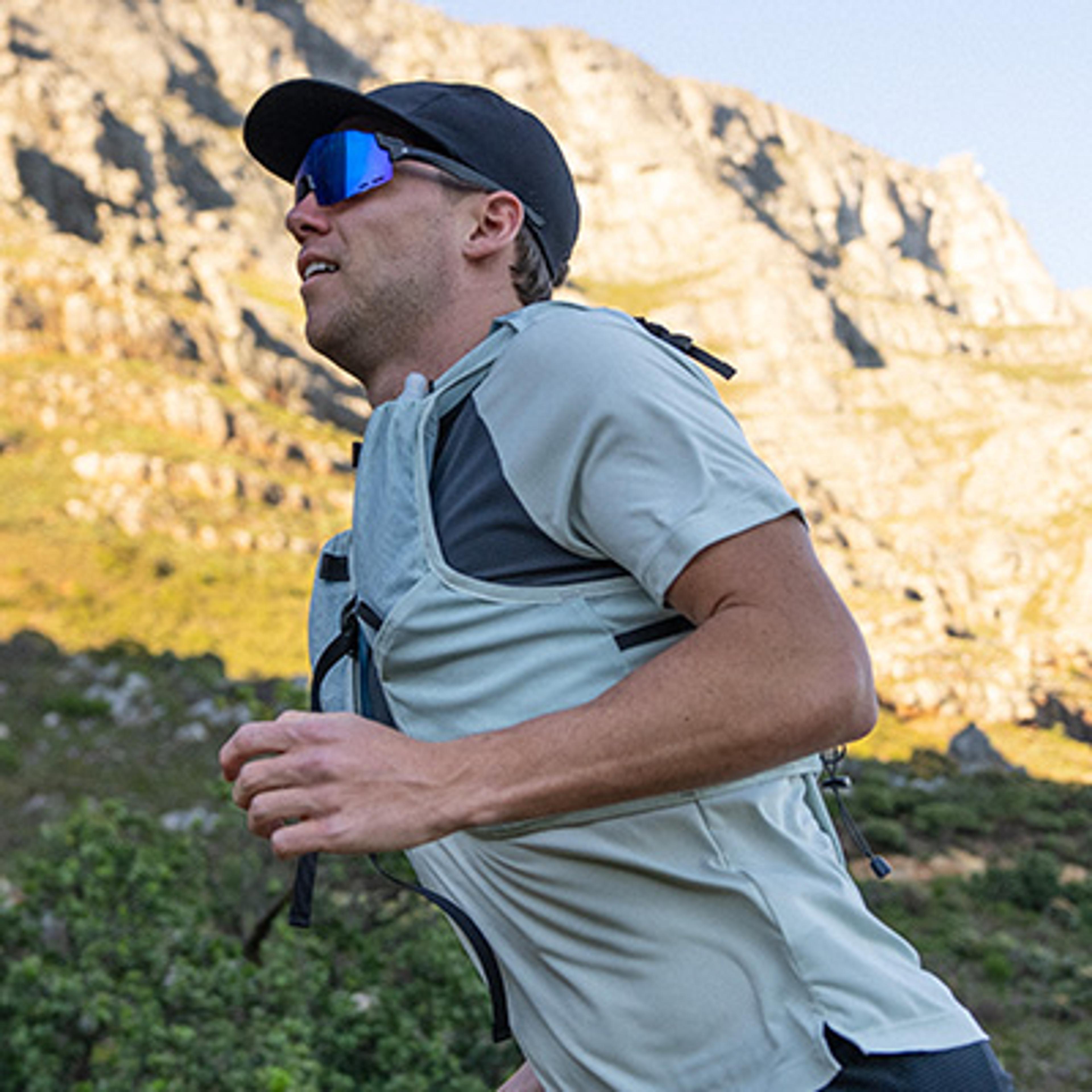 Man in a cap, blue sunglasses, and running vest trail running with mountains in the background.