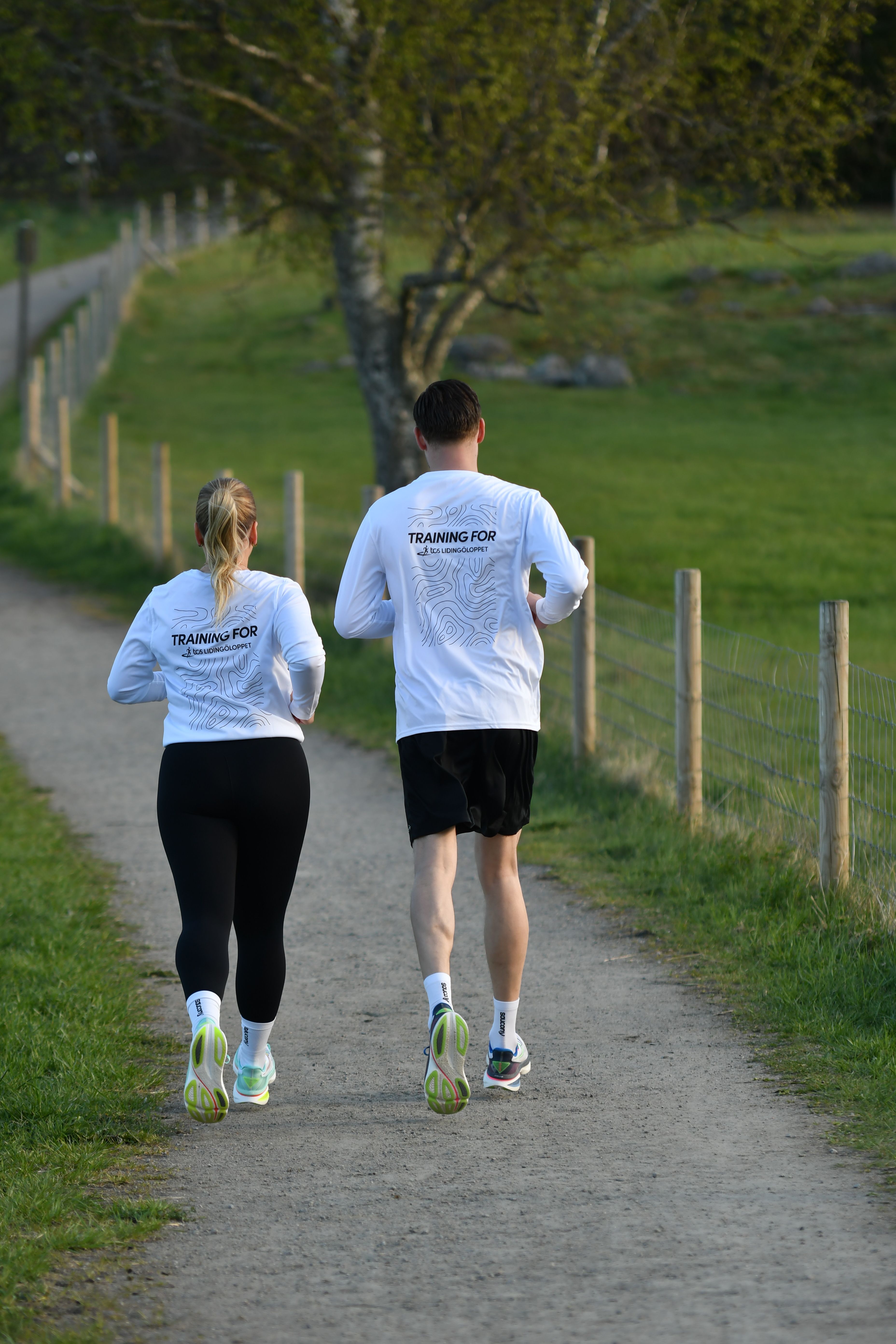 Two people wearing white "TRAINING FOR" shirts jog away on a dirt path through a green, fenced landscape.