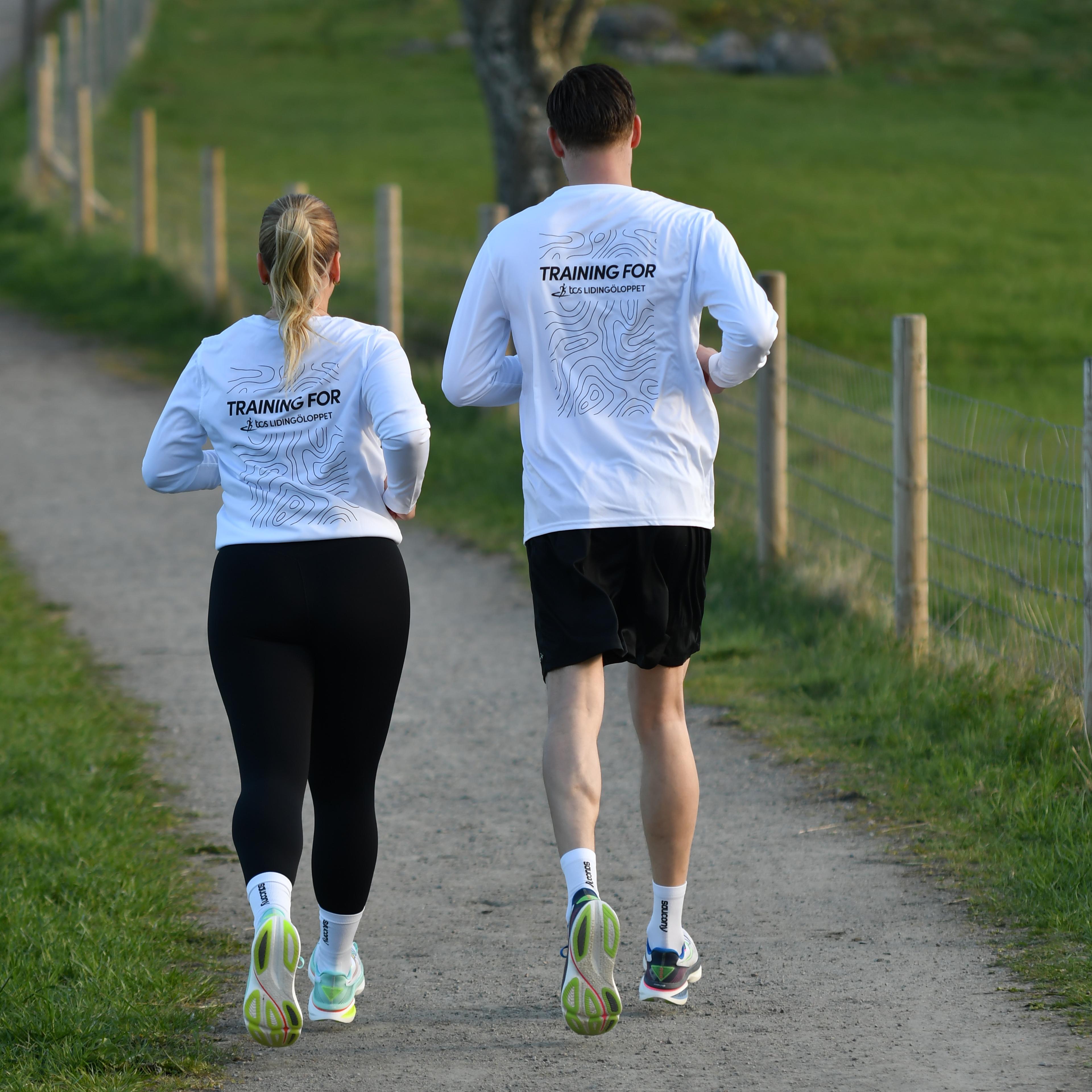 Two people wearing white "TRAINING FOR" shirts jog away on a dirt path through a green, fenced landscape.
