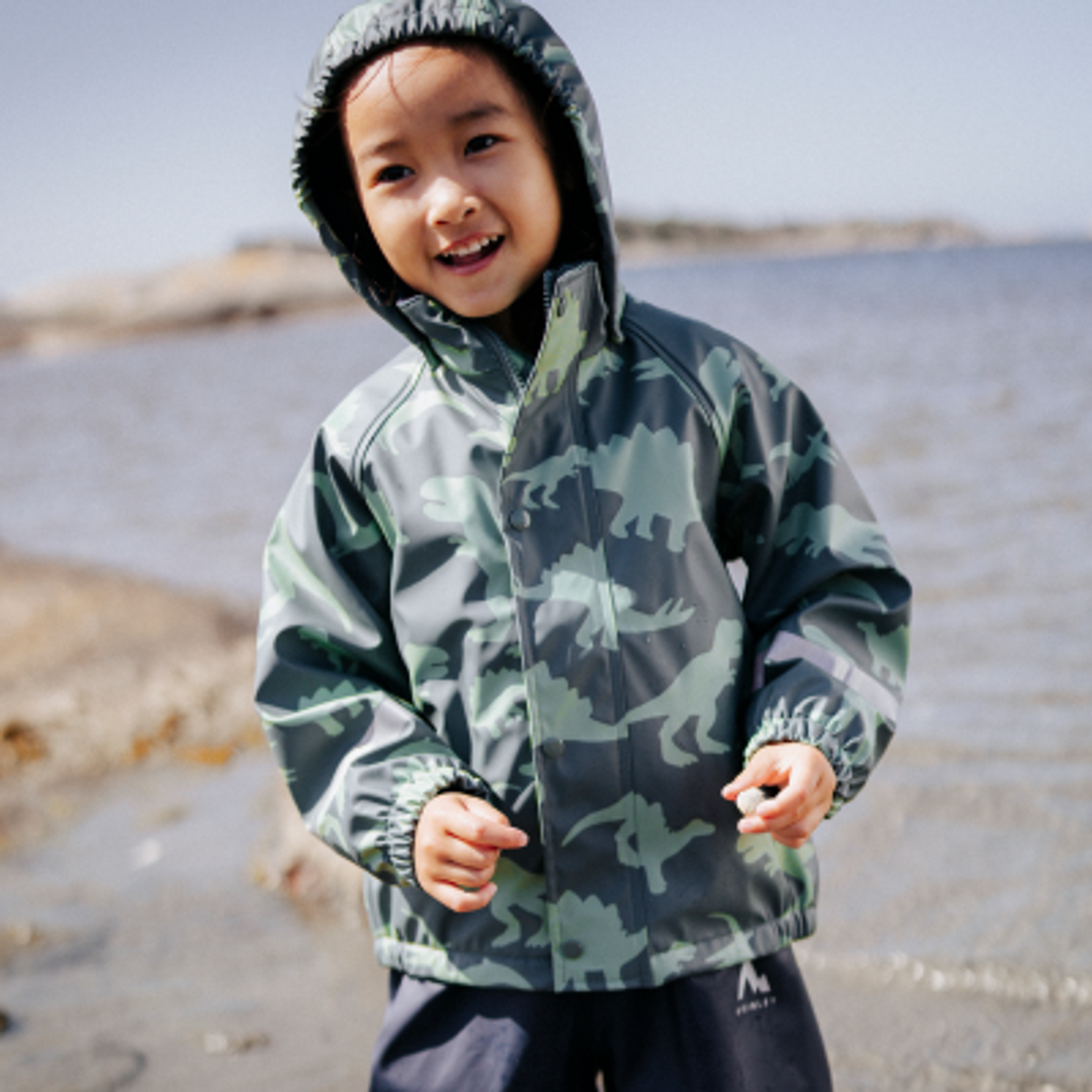 A smiling child wears a hooded green dinosaur rain jacket by the water.
