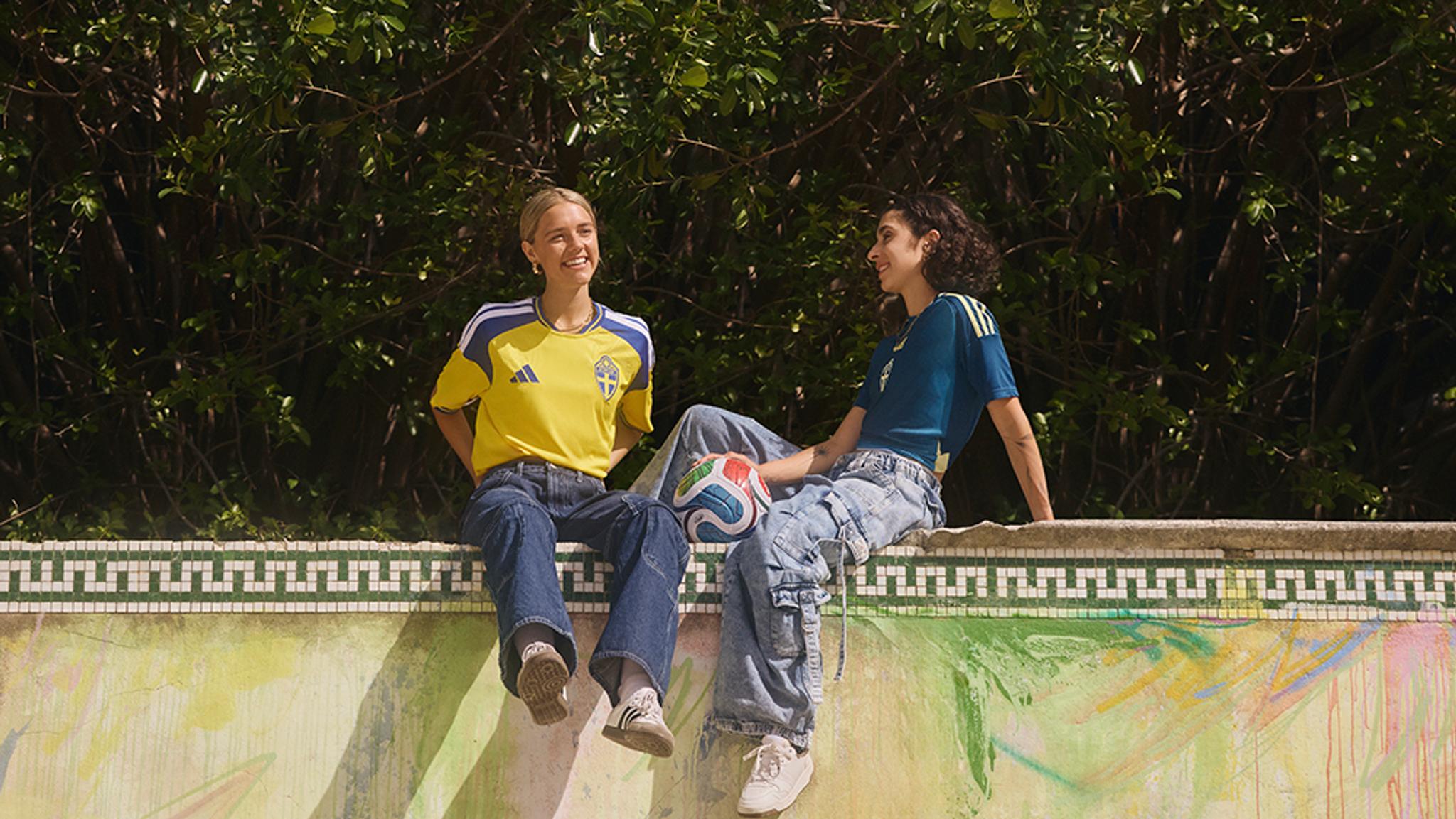 Two women in athletic jerseys sit on a colorful wall, one holding a soccer ball.