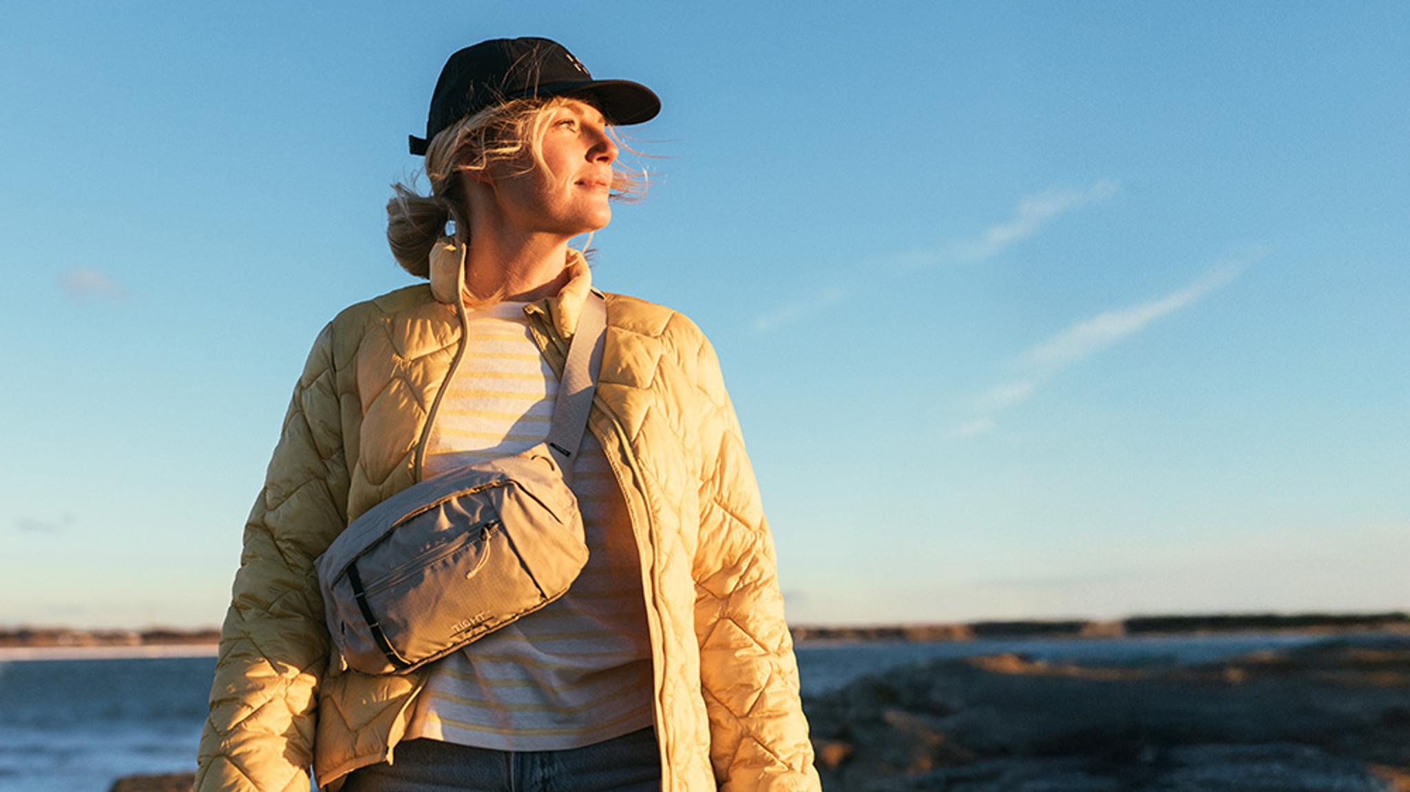 A blonde woman wearing a yellow jacket, black cap, and grey fanny pack looks out over a rocky coast at sunset.